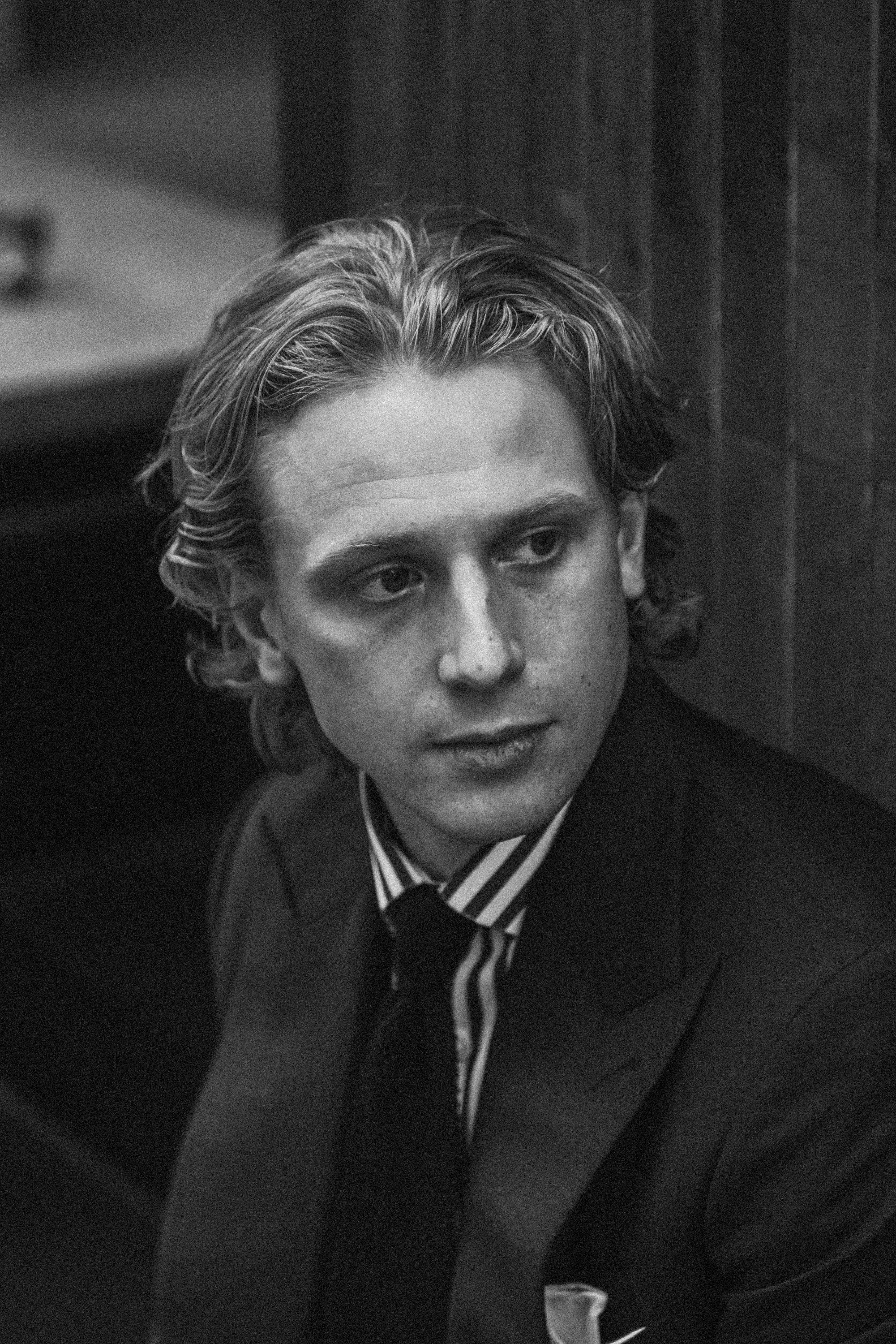 Black and white photo of a man with wavy hair wearing a suit and striped shirt, looking to the side, with a serious expression.