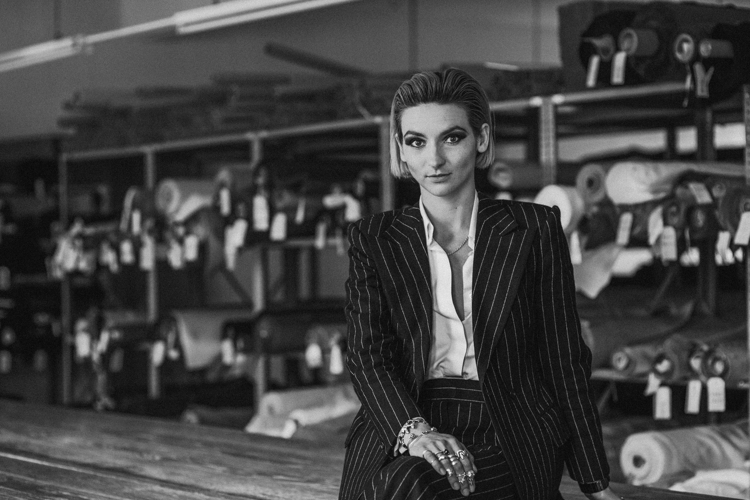 A black and white photo of a woman with short hair, wearing a business suit with pinstripes, standing in front of shelves filled with rolls of fabric or wallpaper, in a warehouse or store setting.