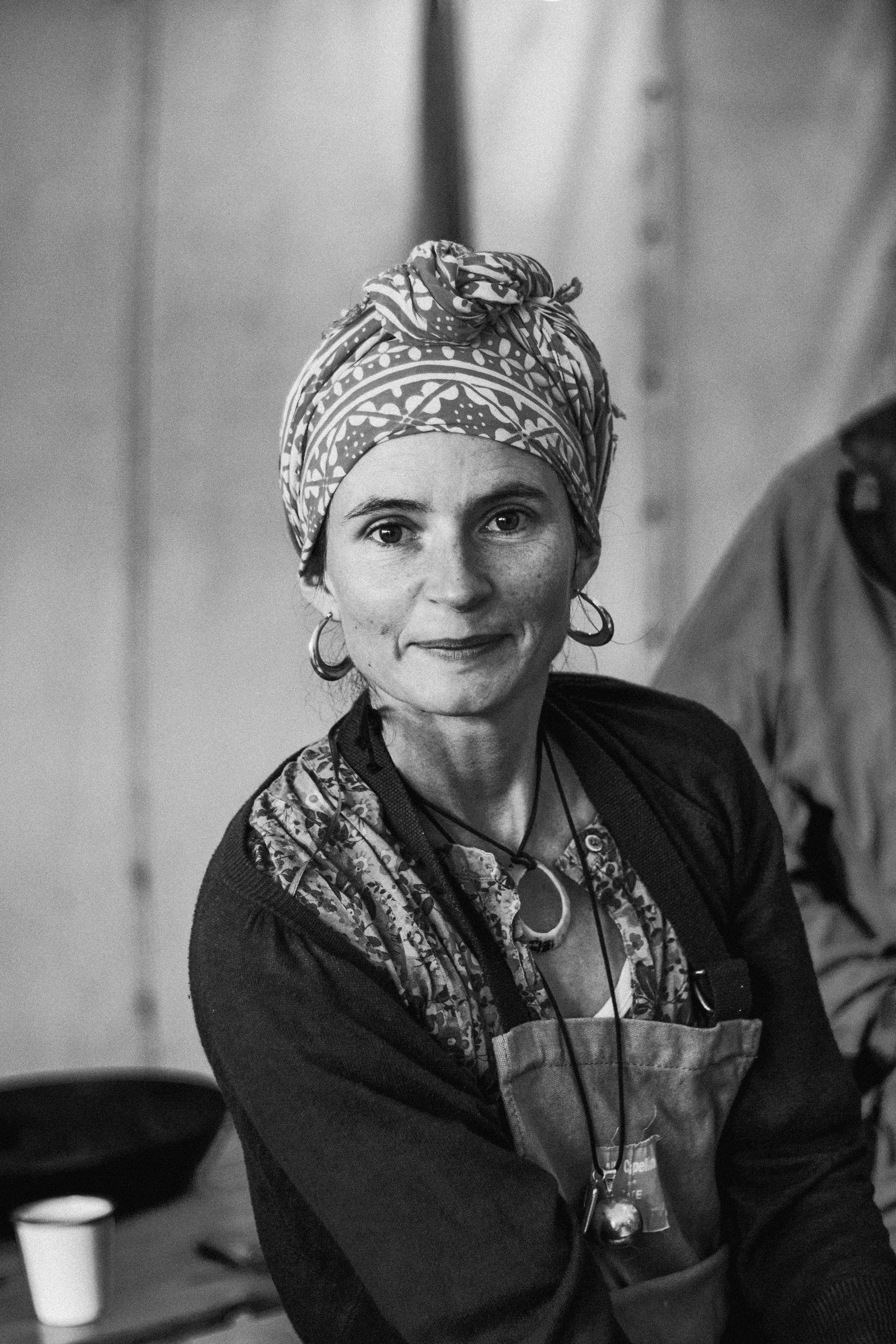 A woman with light skin, freckles, and dark eyes, wearing large hoop earrings and a patterned headscarf, is smiling while looking at the camera. She has short hair underneath the headscarf and is wearing layered necklaces and a dark top with a colorf