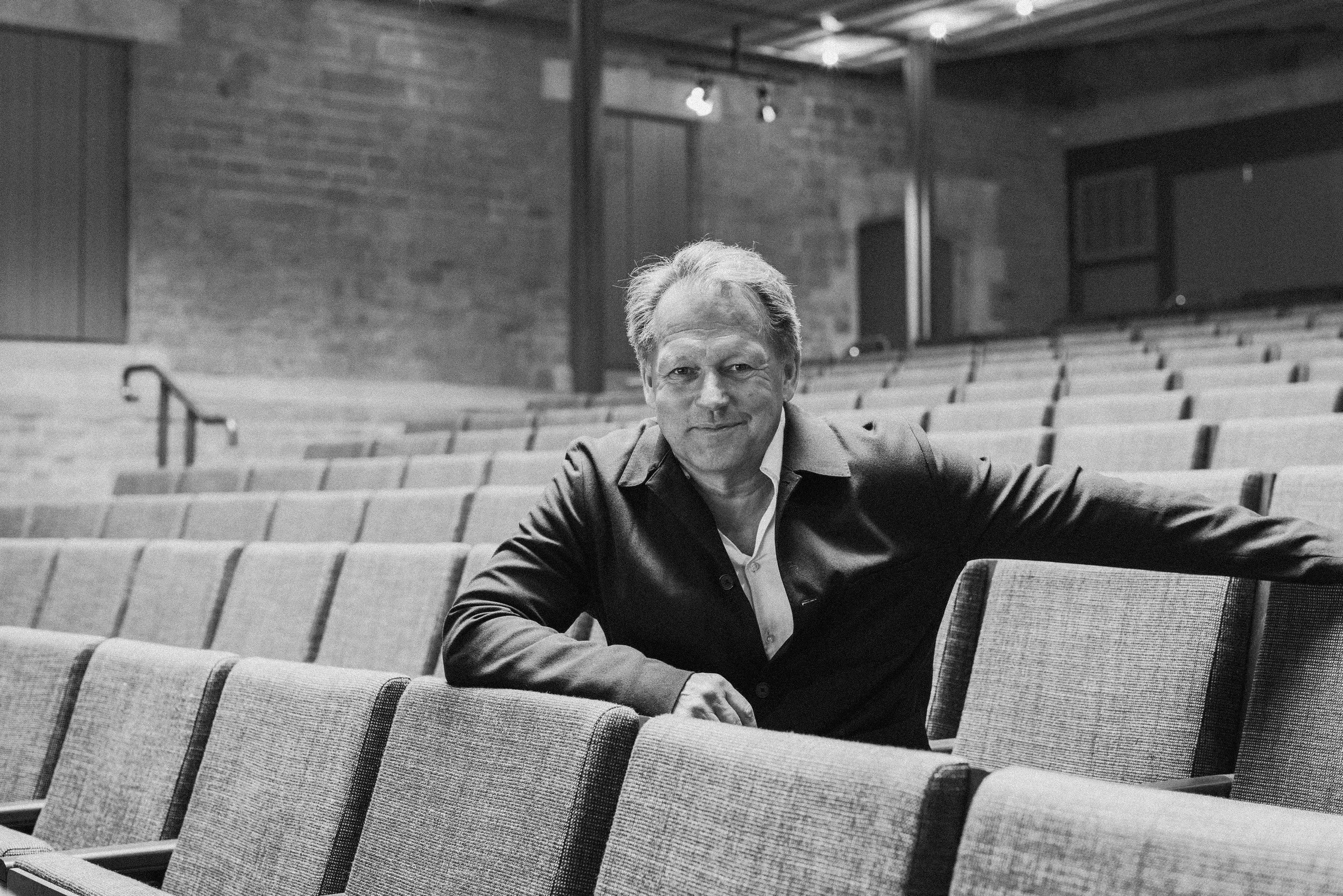 A middle-aged man with gray hair sits in an empty auditorium with rows of empty seats. He is wearing a dark blazer and a light-colored shirt, looking at the camera with a slight smile. The auditorium walls are brick, and there is a television on the 
