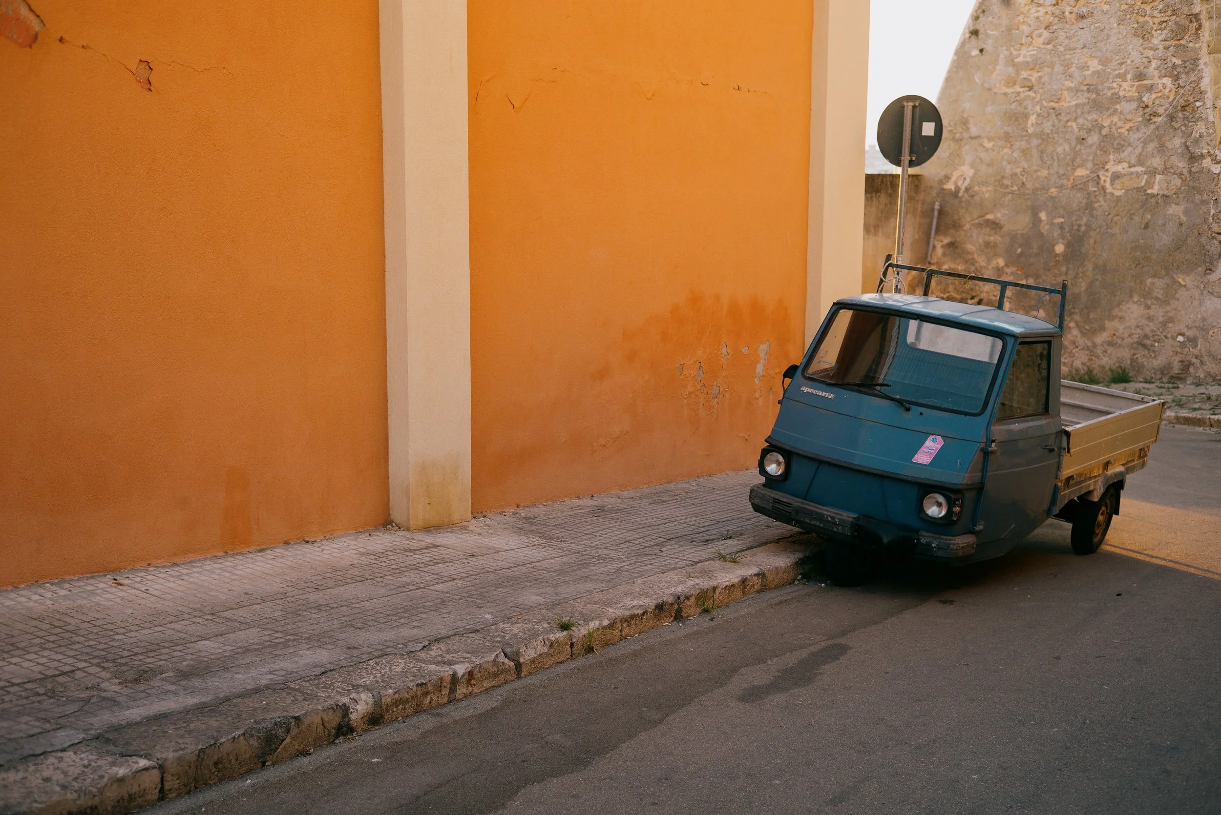 A small blue three-wheeled utility vehicle parked on a sidewalk next to an orange wall with visible cracks and peeling paint, with a stone wall visible in the background in Gallipoli, Italy.
