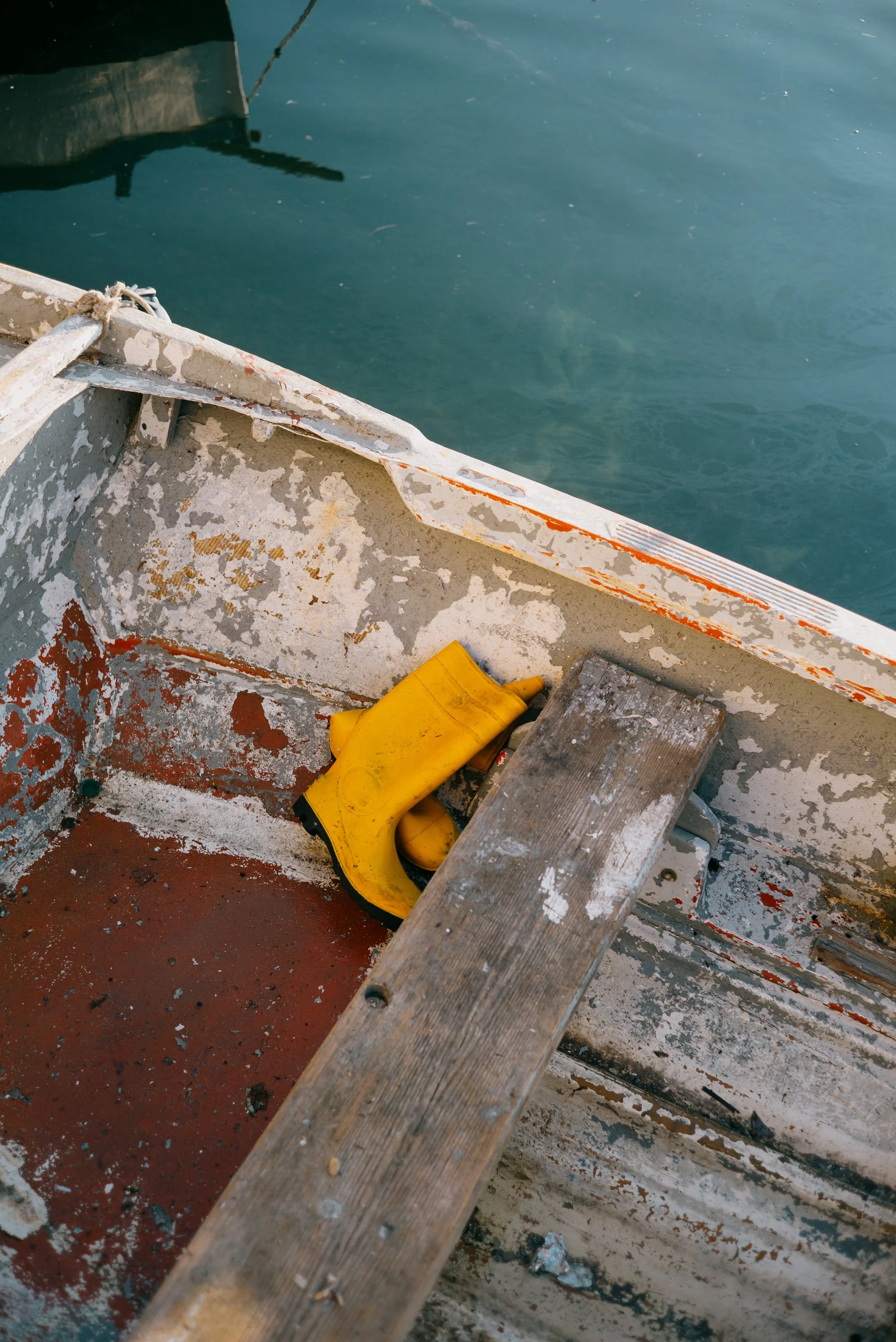 Part of a boat with weathered and chipped paint, a yellow waterproof glove, and a small wooden block on the worn floor with peeling paint, next to calm blue water in Gallipoli.