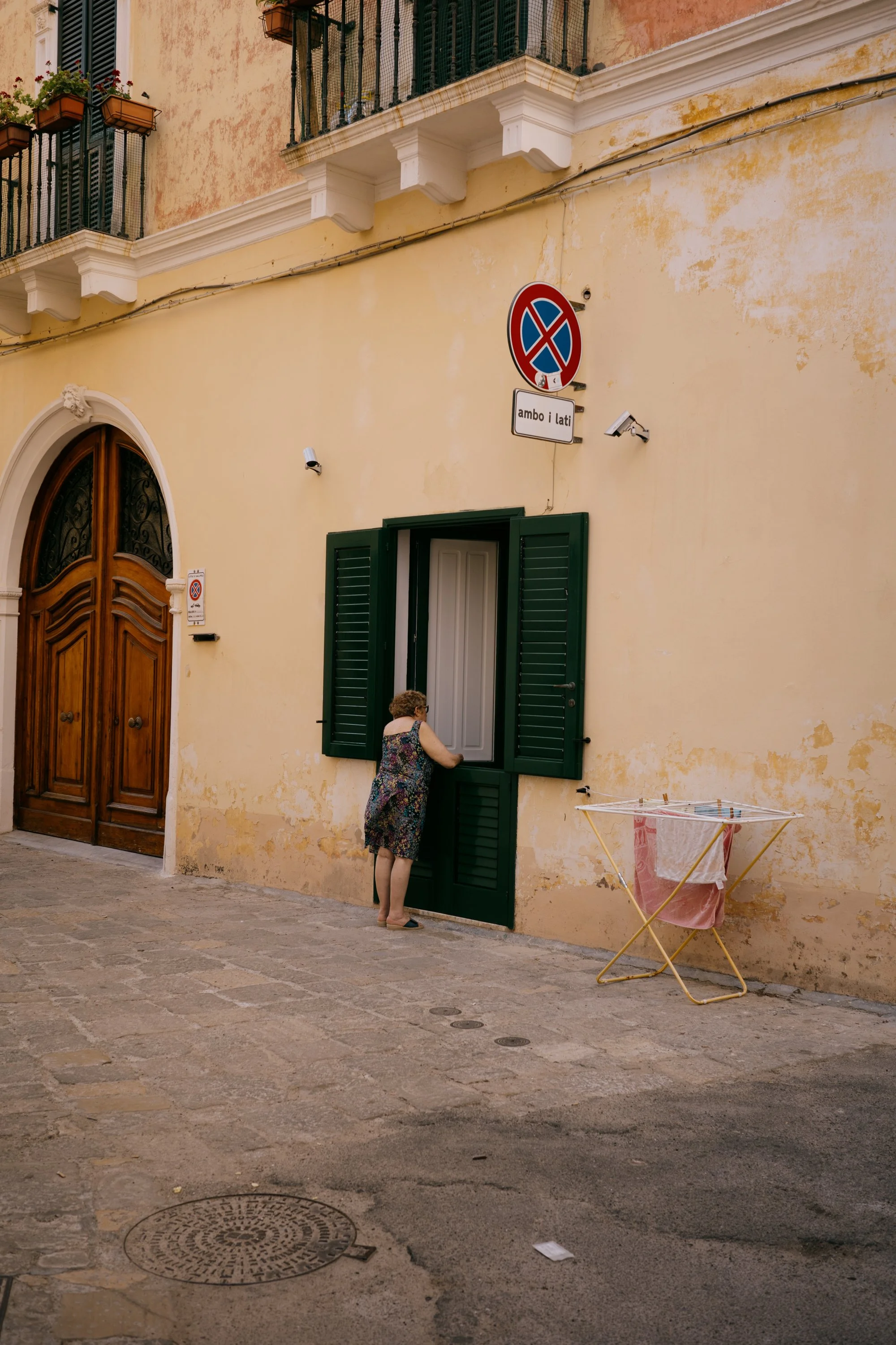 A woman with short curly hair wearing a floral dress and black shoes, stands on a cobblestone street next to a large, dark green window with open shutters in a yellow building. The building has a wooden arched door, potted plants with red flowers on 
