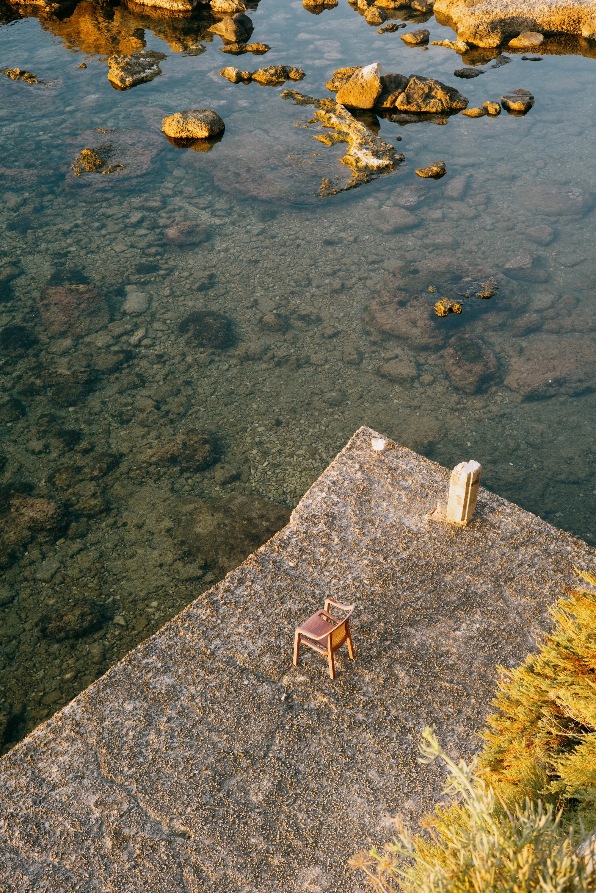 A solitary chair on a concrete platform near a body of water with rocks, some covered in moss, and sparse vegetation in the foreground.
