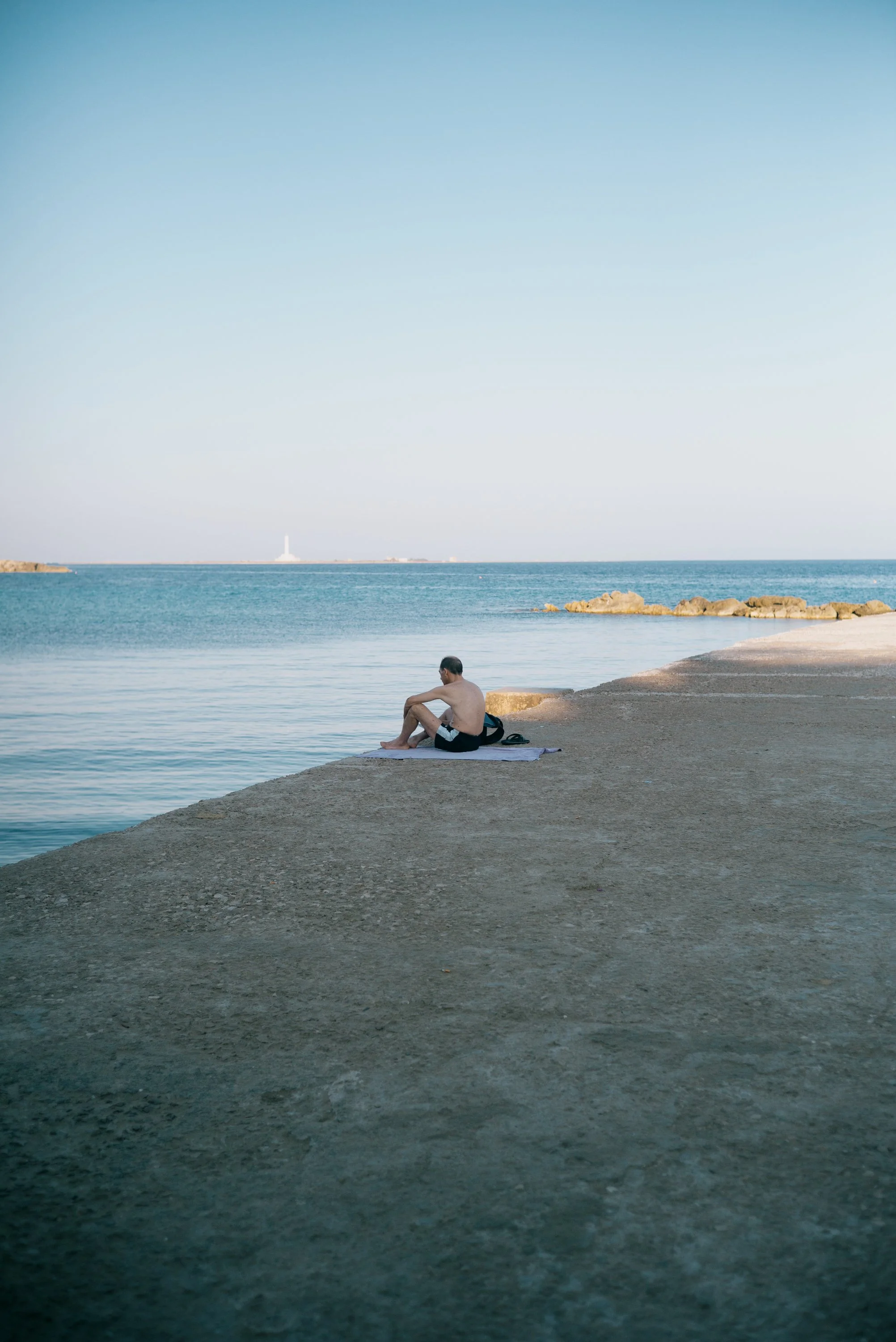 A man sits on a towel on a concrete pier at the beach, looking out at the ocean with rocks and a lighthouse in the distance under a clear blue sky in Italy;