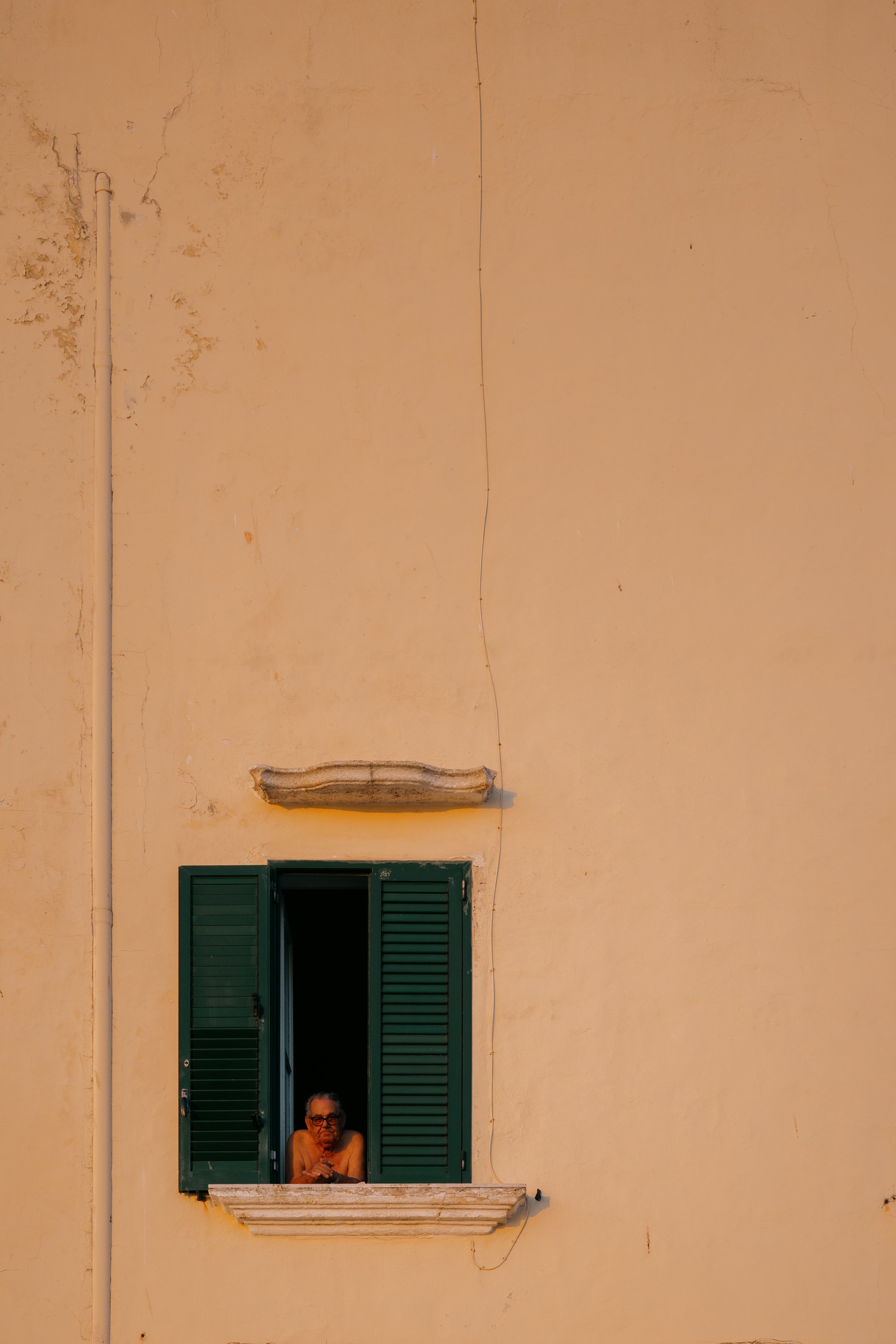 An elderly man with glasses sitting at an open window with green shutters on a yellowish building wall in Italy. 