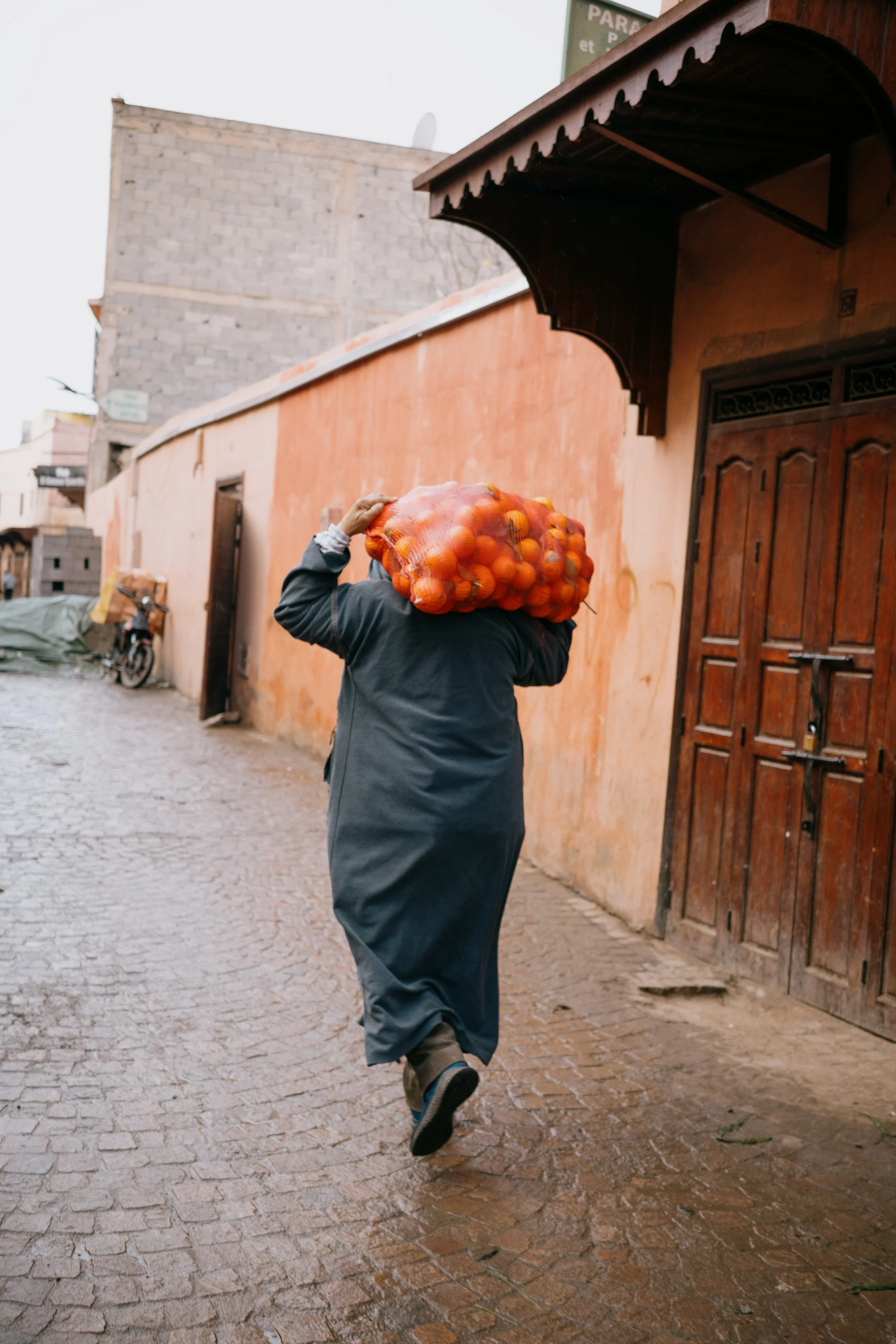 A person walking on a cobblestone street carrying a large bag of oranges on their shoulder in an urban area.