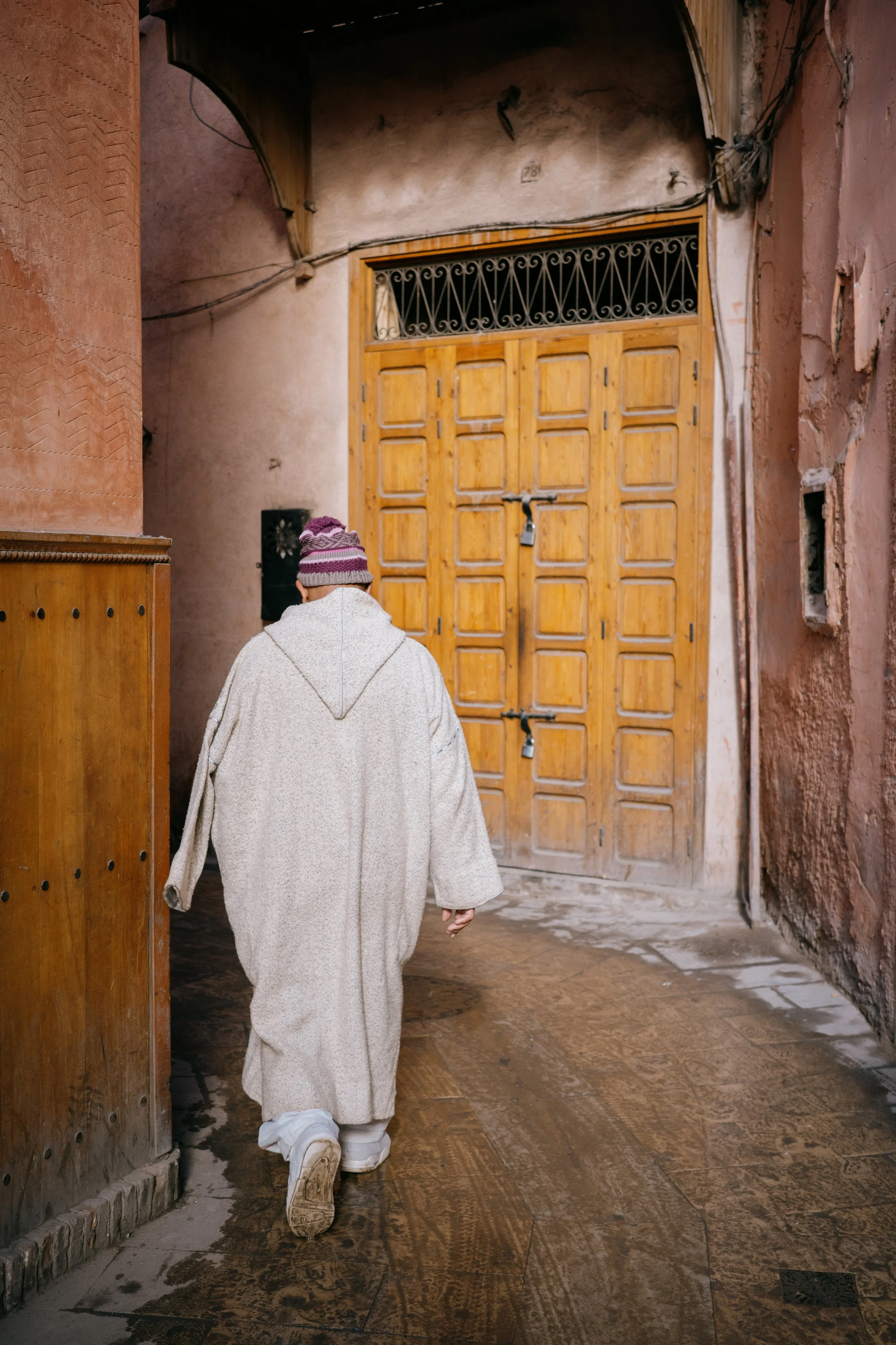Person wearing a gray hoodie, white pants, and a purple-striped knit cap walking towards a wooden door in the streets of Marrakech