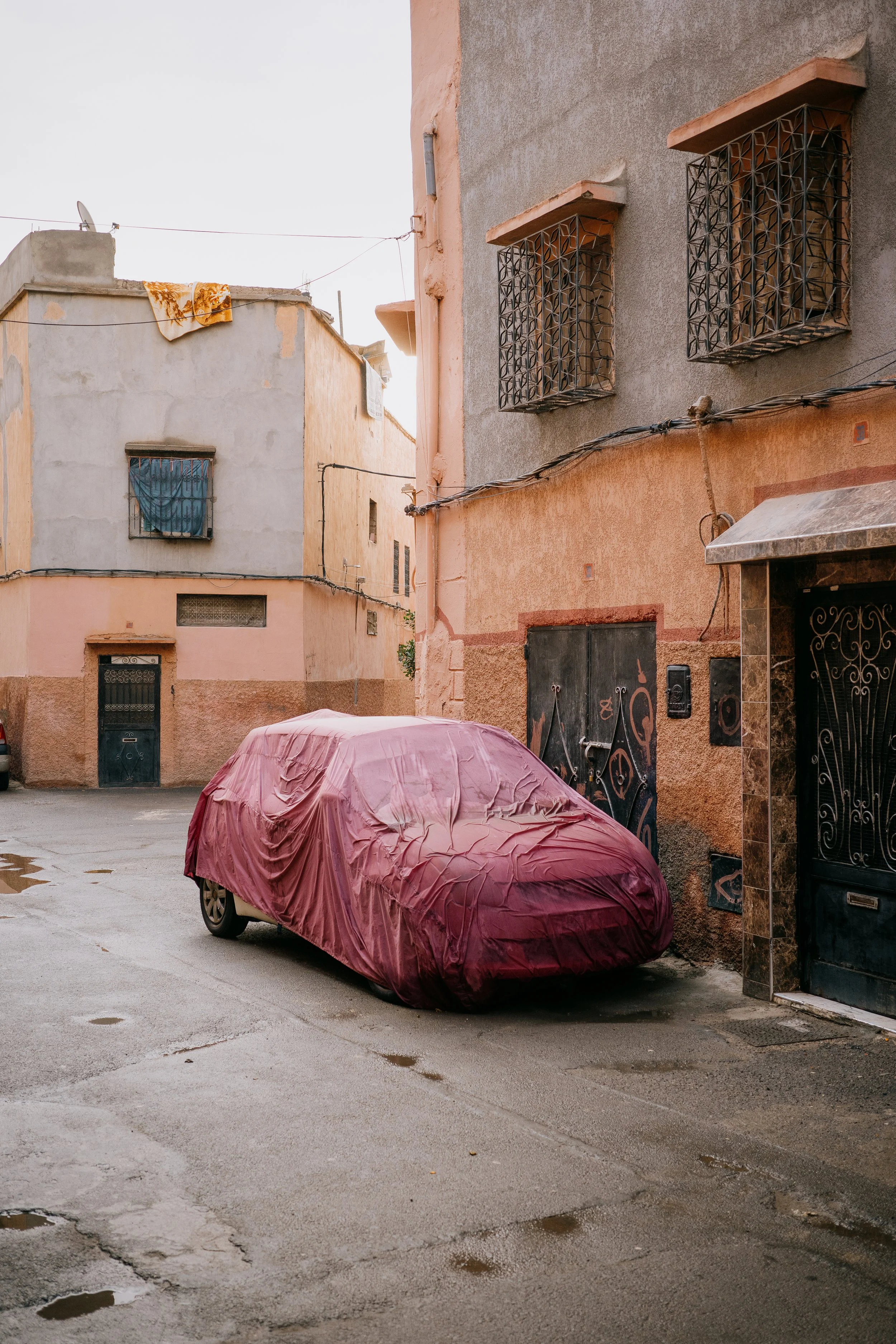 An urban scene with a parked car covered by a burgundy protective cover, wet pavement with puddles, and buildings with barred windows, some with cloth hanging, under an overcast sky.