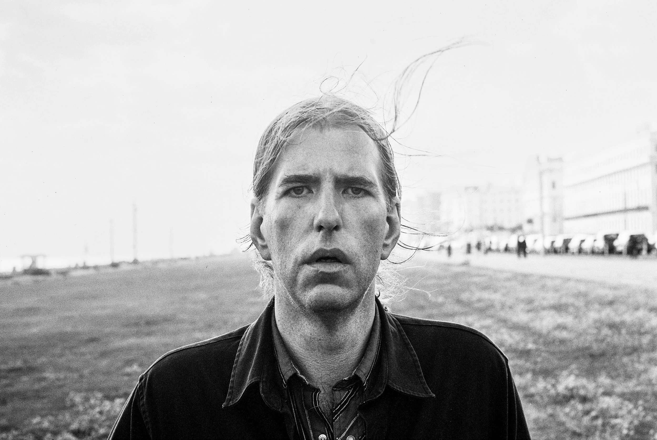 A black-and-white portrait of a man with long hair standing outdoors near a body of water, with buildings in the background.