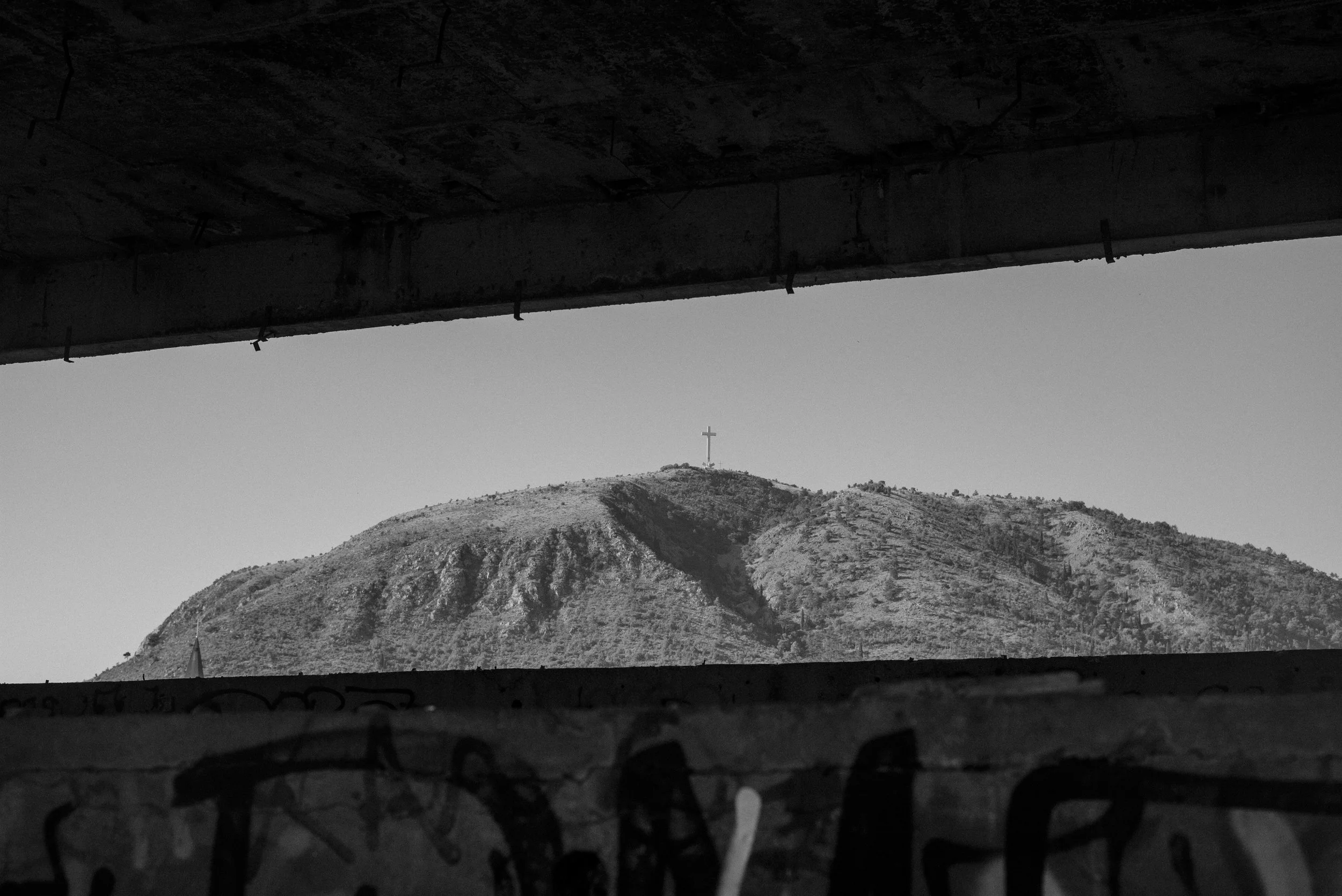 View of a mountain with a cross on top, seen through a partially open window with bars in Mostar, Bosnia. 