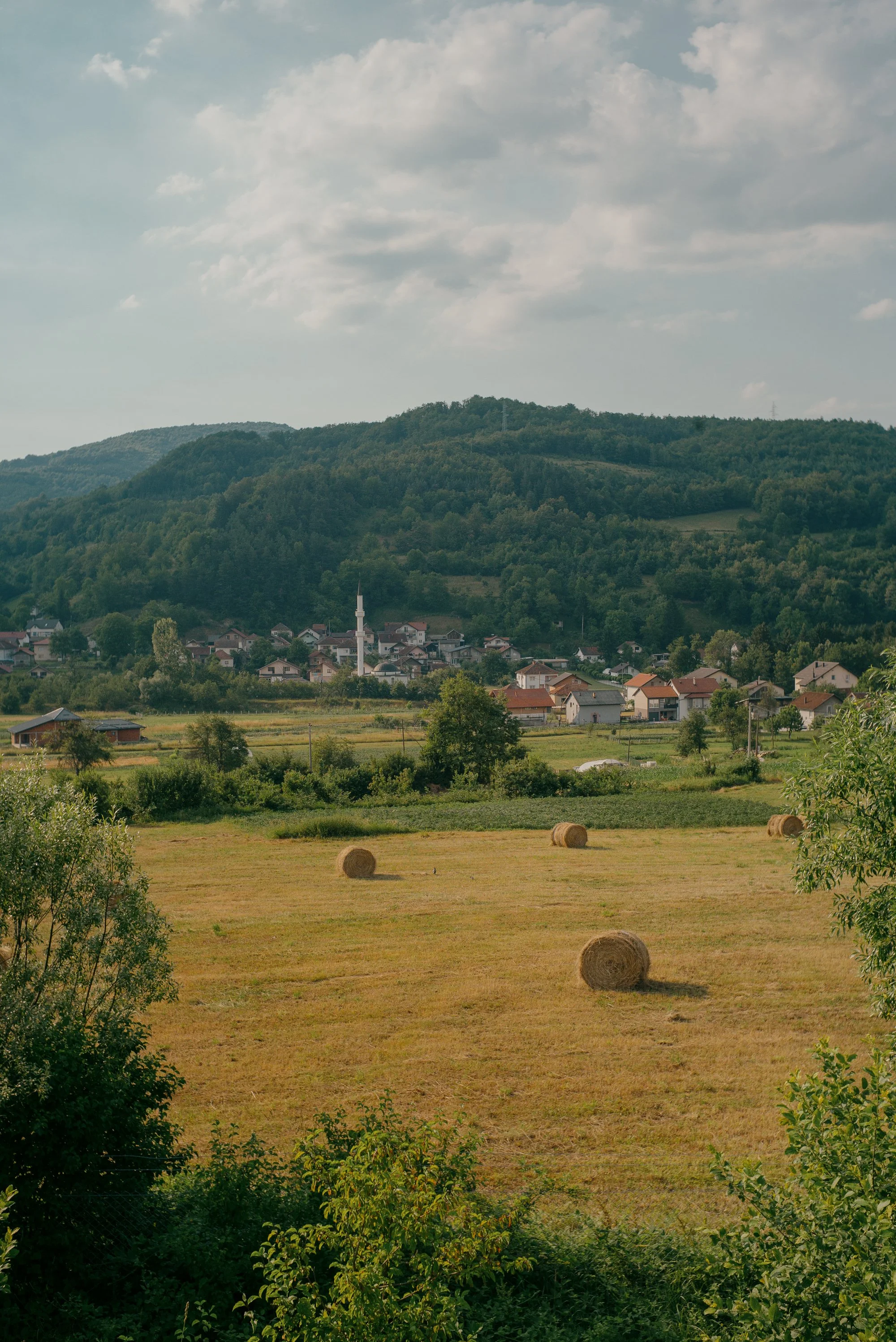 A rural landscape with hay bales in a field in the foreground, a small village with houses and a church in the middle ground, and forested hills in the background under a partly cloudy sky.