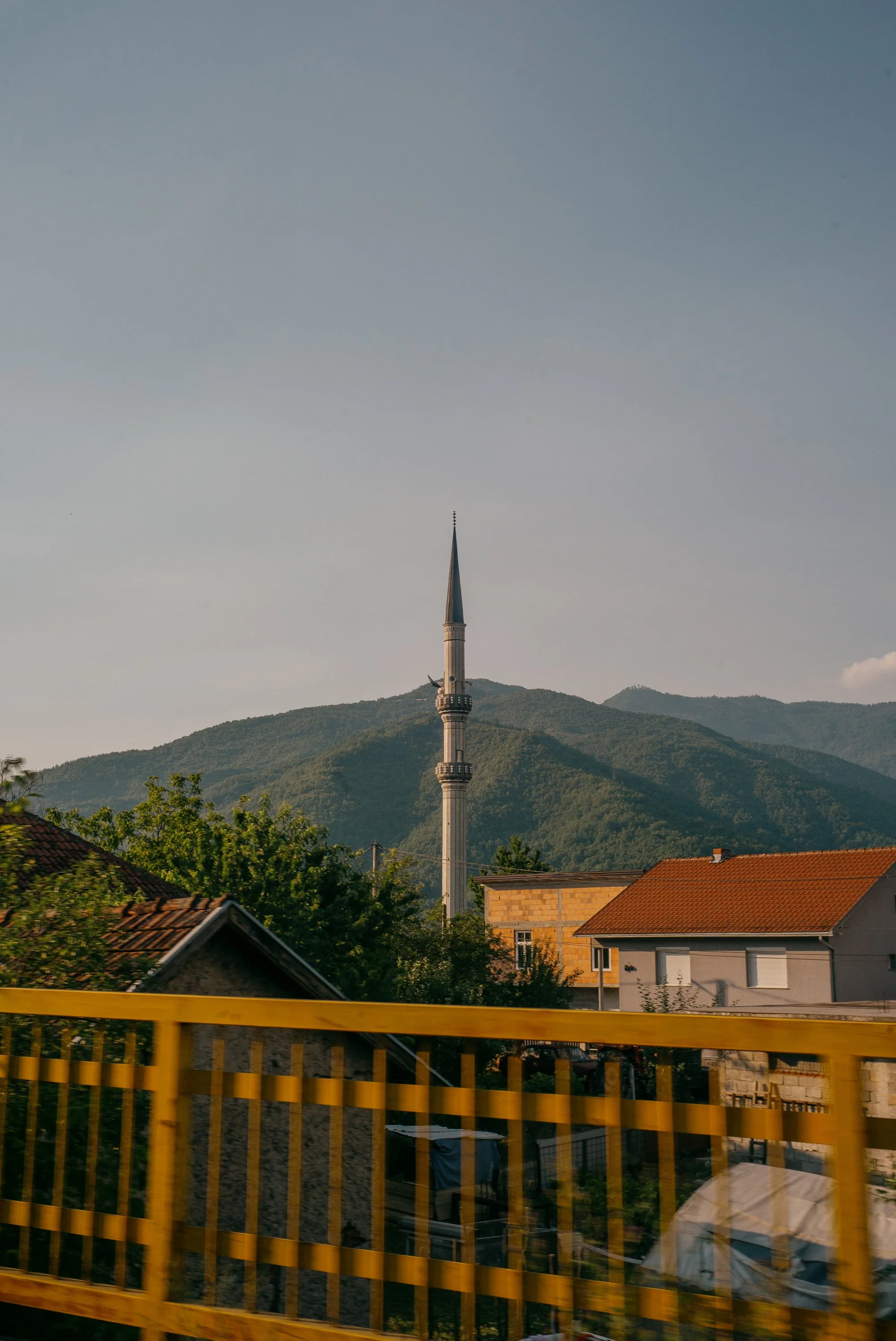 Town with houses, trees, and a tall minaret with a blue spire against a mountain landscape and a cloudy sky. Bosnia. 