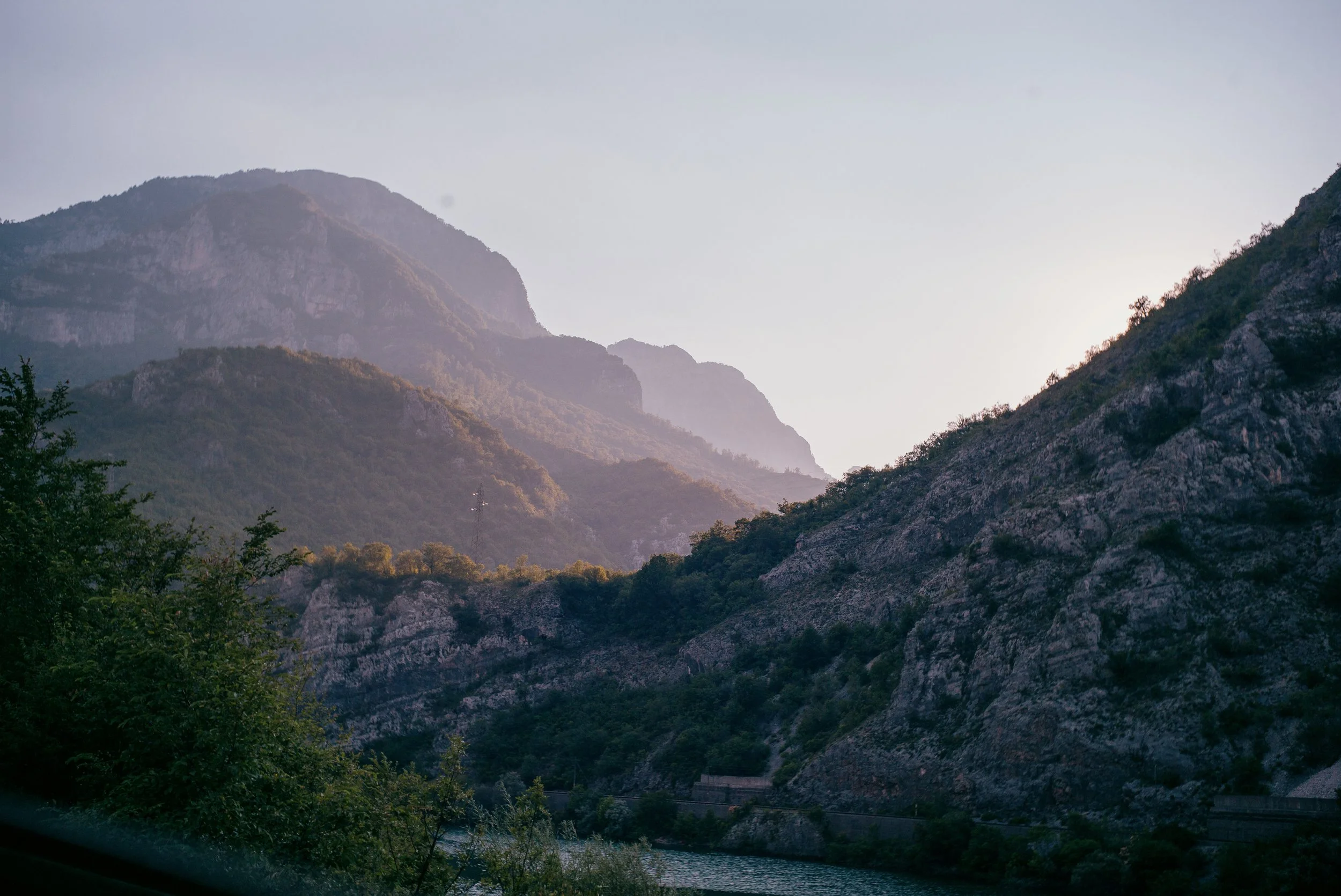 A scenic landscape of mountains with lush green vegetation and rocky cliffs, under a clear sky, with a river flowing at the bottom.