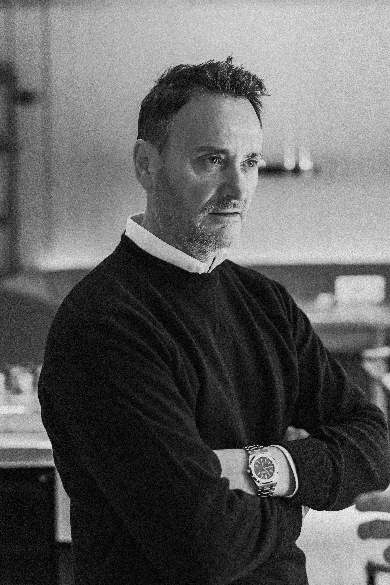 A black-and-white photo of a middle-aged man with a beard and styled hair, wearing a dark sweater with a white shirt collar underneath, standing with crossed arms, and looking to the side. He is wearing a wristwatch on his left wrist, indoors with a 