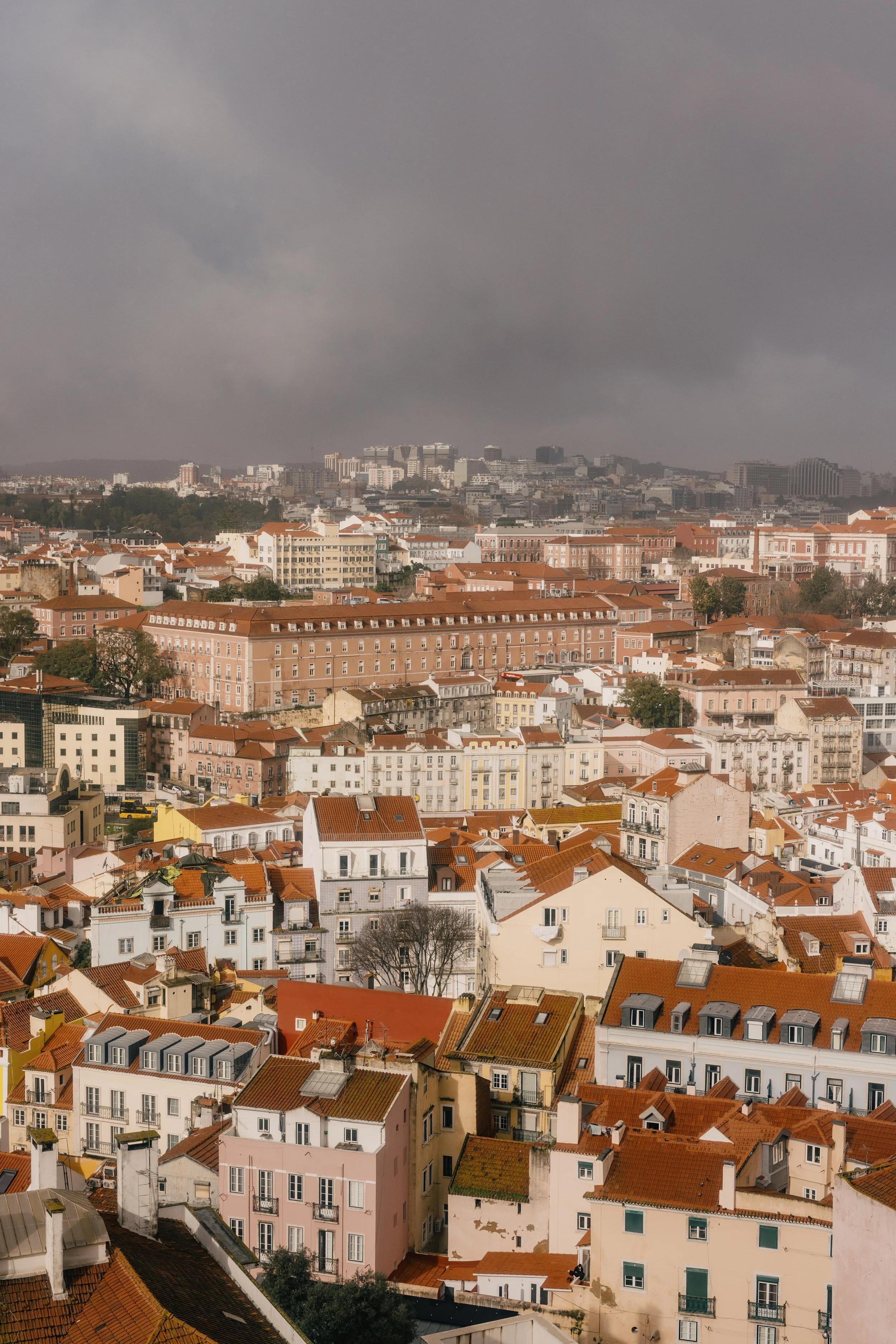 Cityscape with dense buildings, mostly white and pastel-colored, with many red-tiled roofs under a dark, cloudy sky.