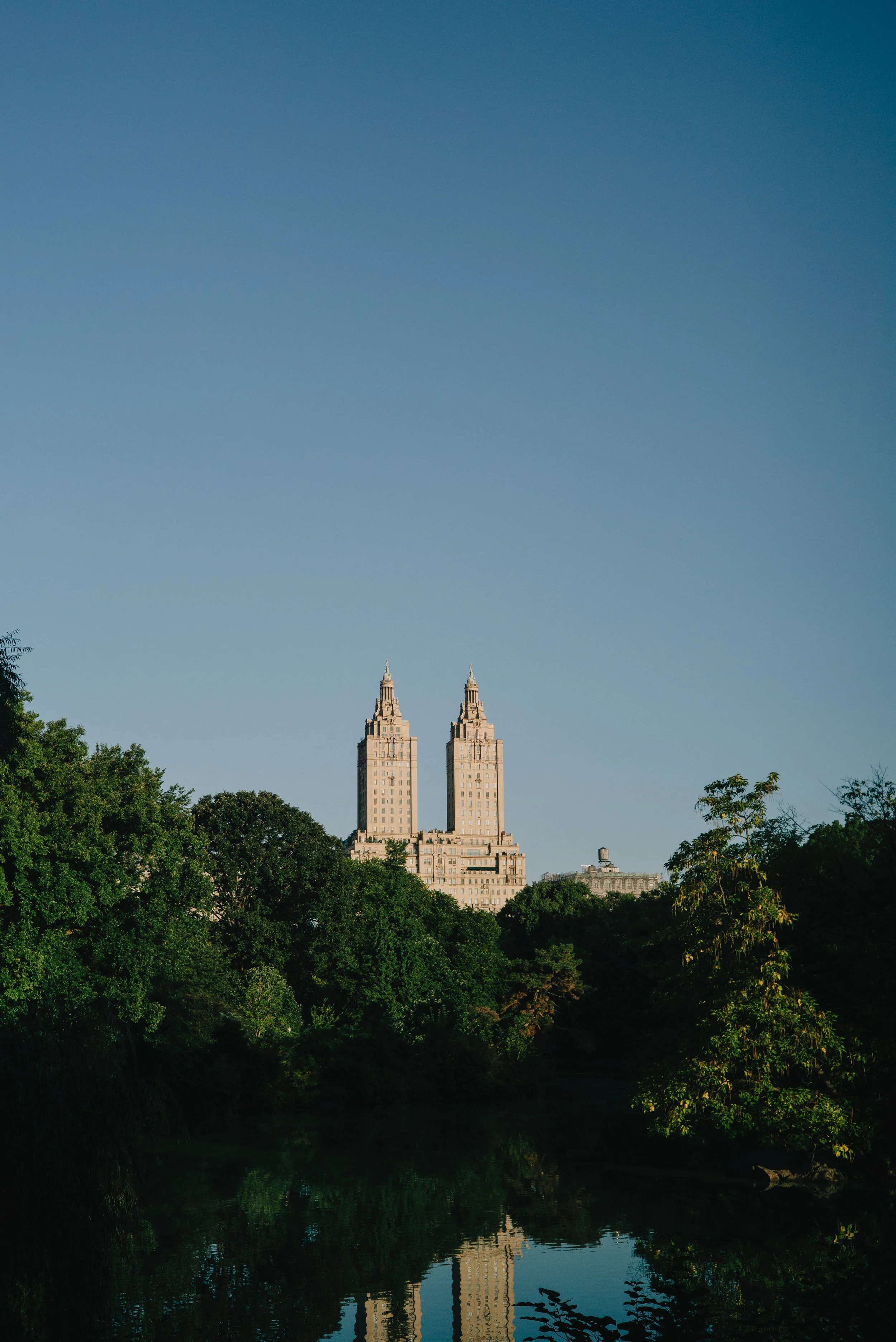 A view of tall twin buildings with a clear blue sky, surrounded by green trees and a small body of water in the foreground.