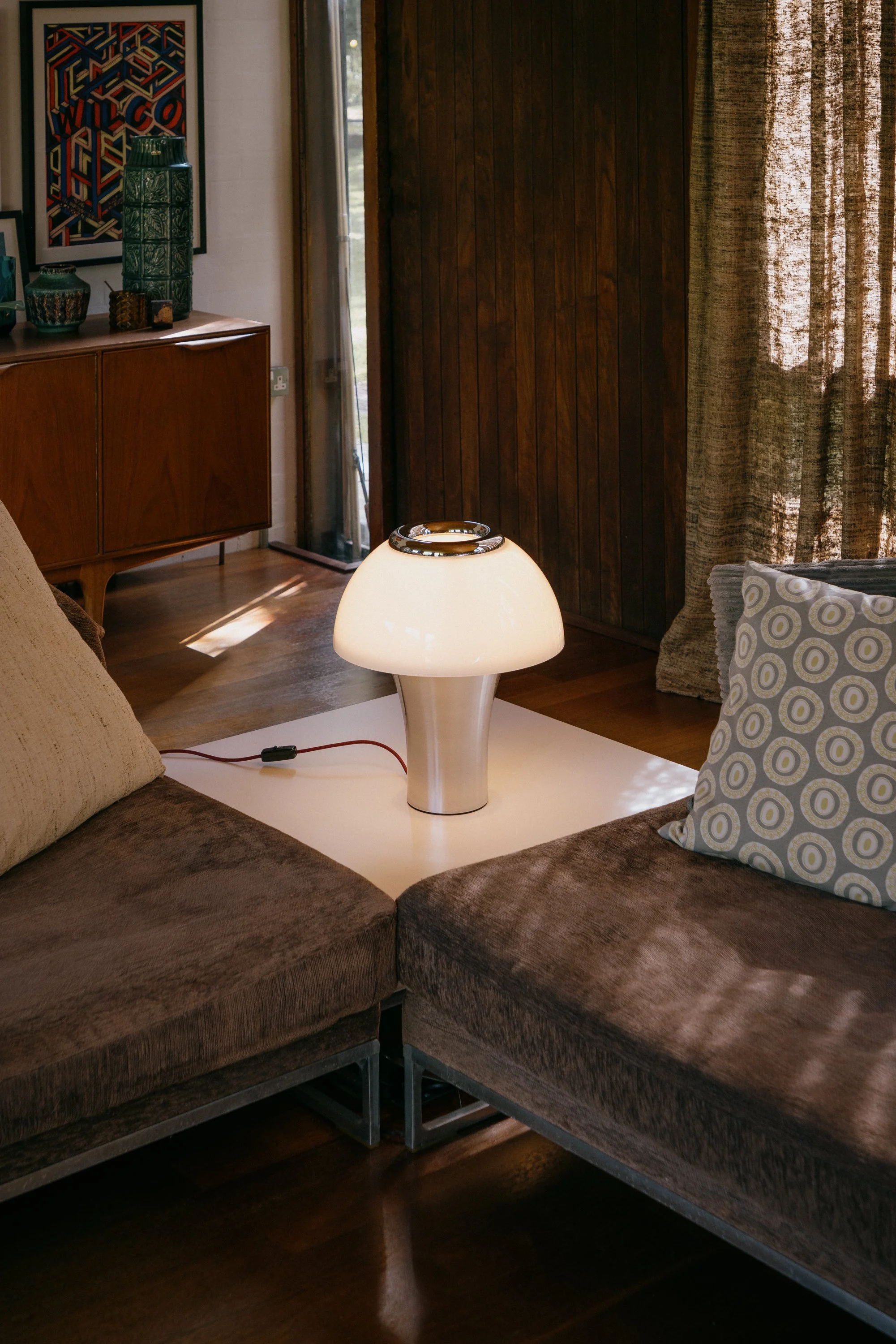 Living room with brown sofa, decorative pillows, a white modern table lamp, wooden cabinet on the wall with green vases, framed artwork, curtains, and a door to the outside.