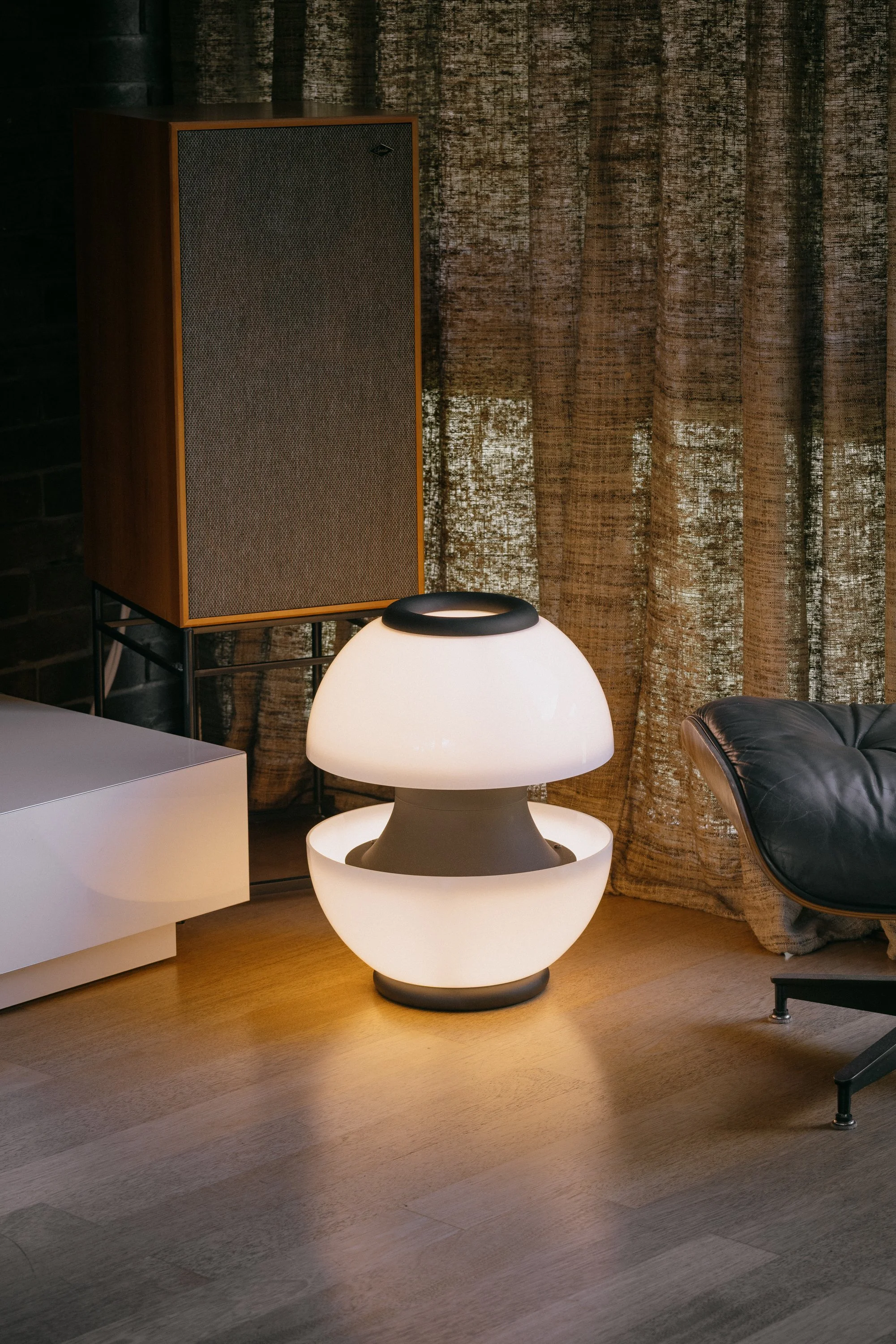 Modern living room with a large, rounded white lamp on a wooden floor, a black leather chair, a white side table, and wooden curtains in the background.