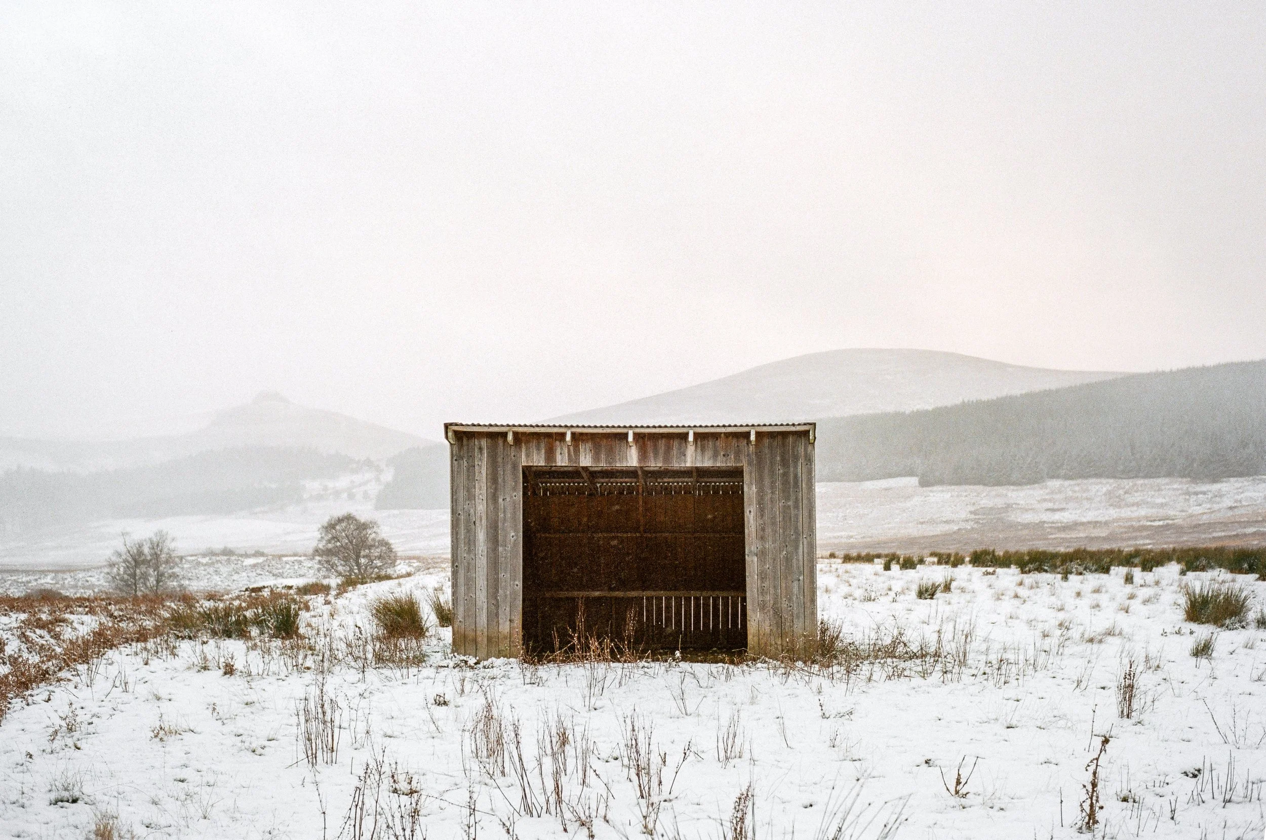 A small wooden shed or barn in an open snowy landscape with hills and sparse trees in the background in Scotland. 