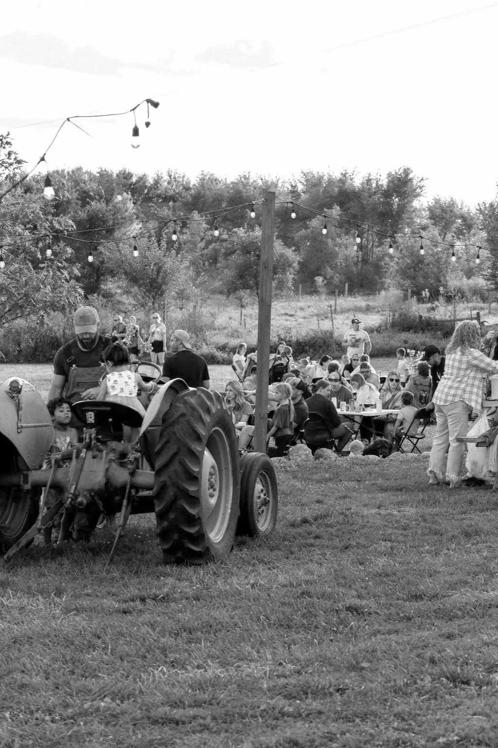 An outdoor gathering with people sitting at tables, a tractor in the foreground, and string lights overhead, suggesting a festive event in a rural setting.