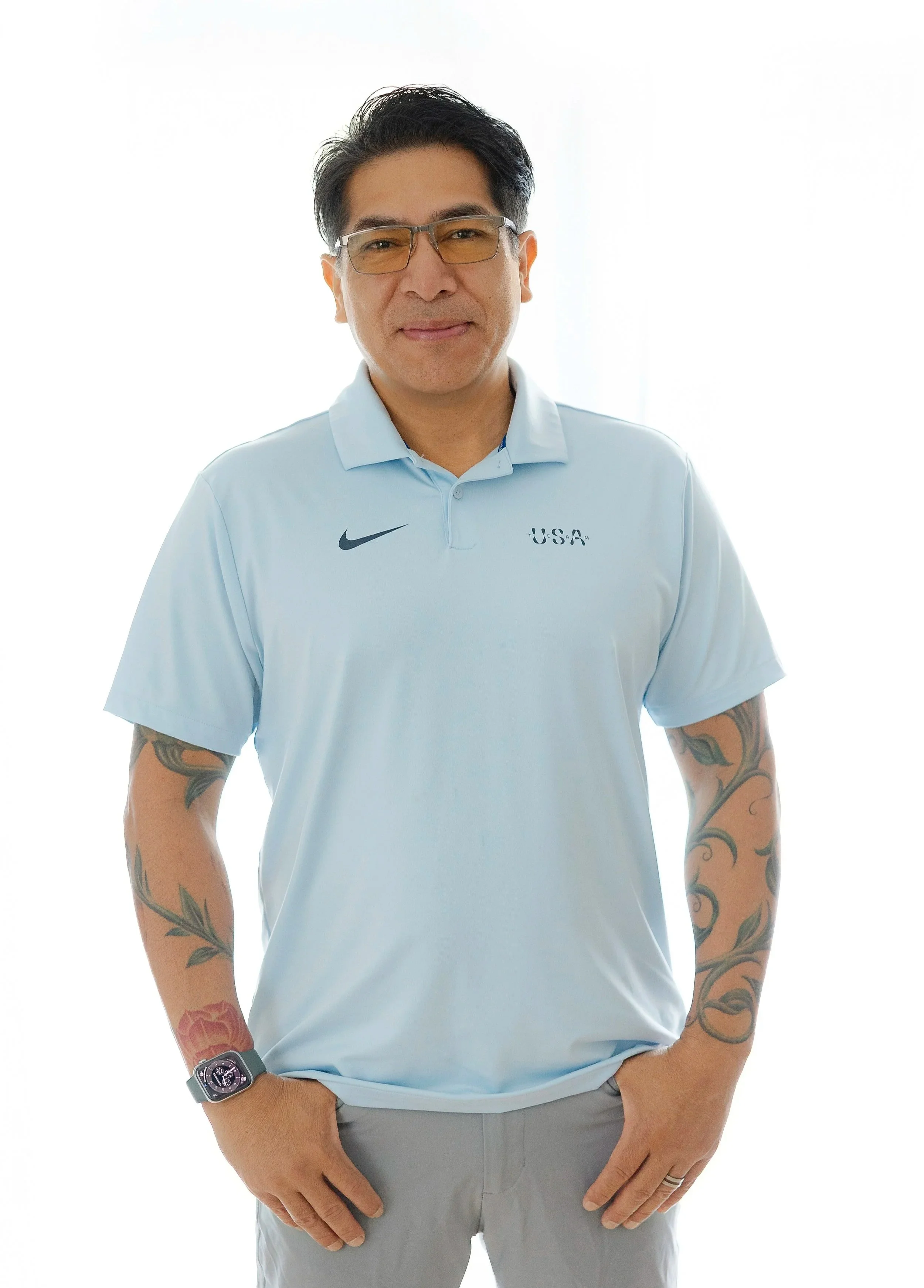 A man with short dark hair, glasses with yellow lenses, and tattoos on his arms, wearing a light blue Nike polo shirt with 'USA' embroidery on the chest. He is smiling slightly, with one hand resting on his hip and the other hanging by his side, against a plain white background.