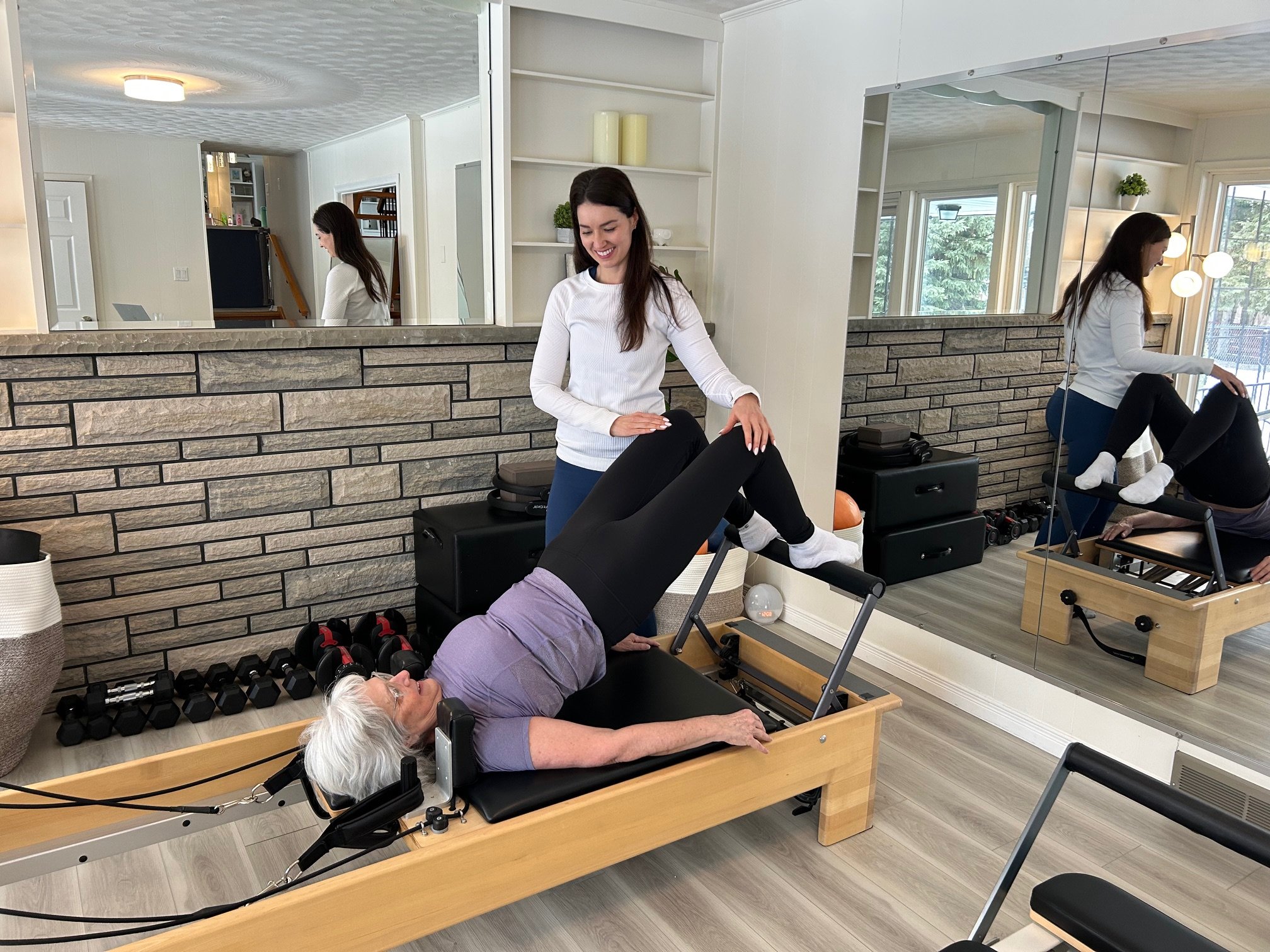 A woman is lying on a Pilates reformer machine while an instructor assists her with a leg stretch. The scene is in a well-lit exercise studio with a mirror, shelves, and gym equipment.