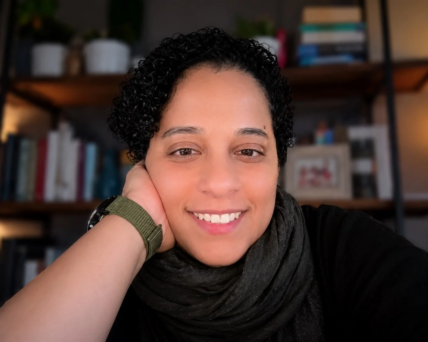 A woman with short curly dark hair, smiling and resting her head on her hand, wearing a green watch and black scarf, in front of a bookshelf.