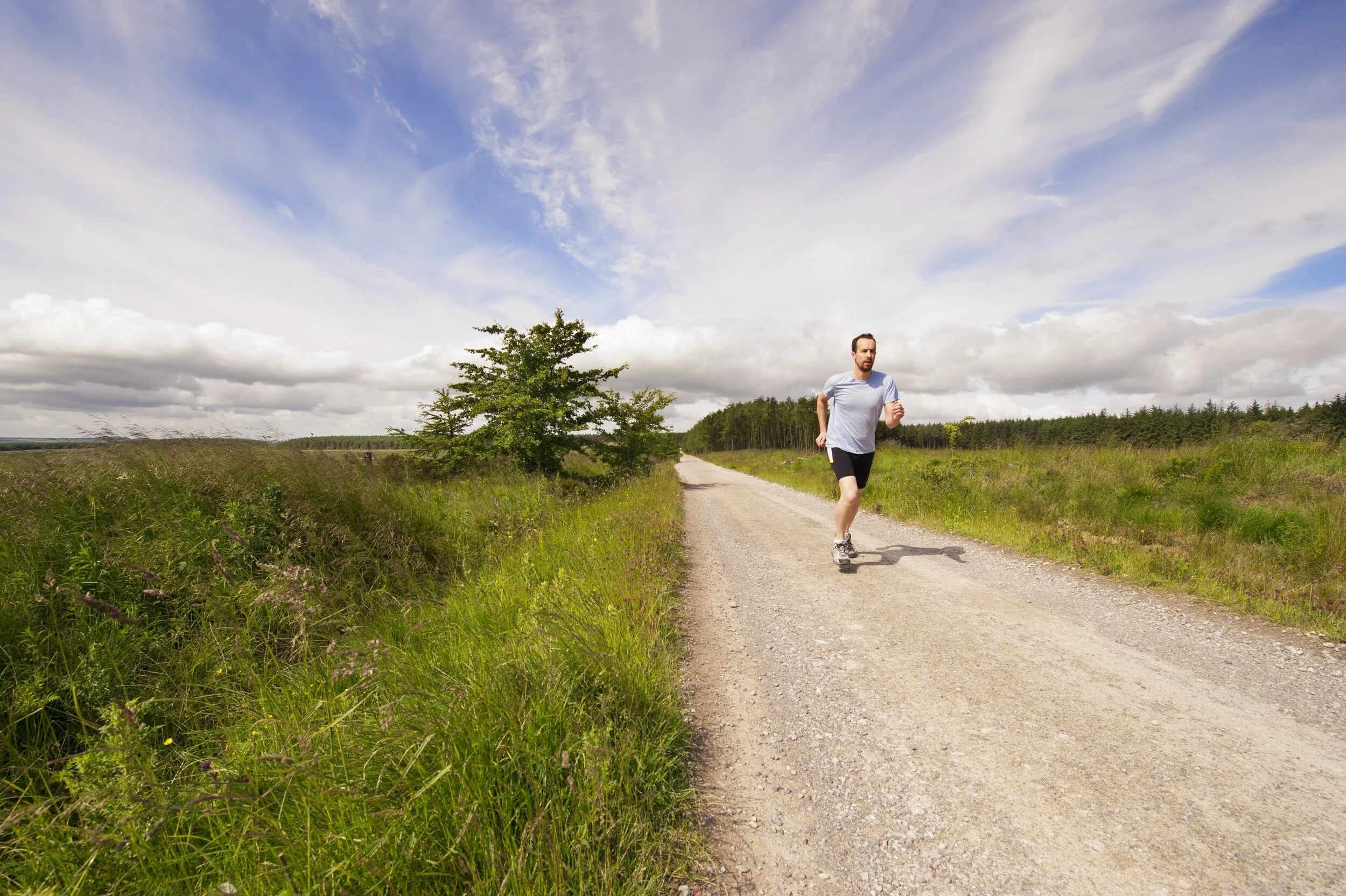 A man jogging on a dirt path surrounded by green fields and trees under a partly cloudy sky.