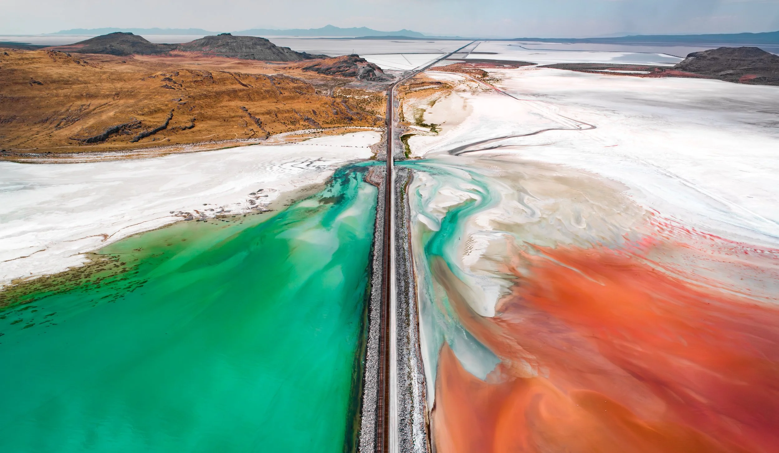 Aerial view of a landscape with a railway track running through colorful salt flats and mineral ponds, with mountains in the distance.