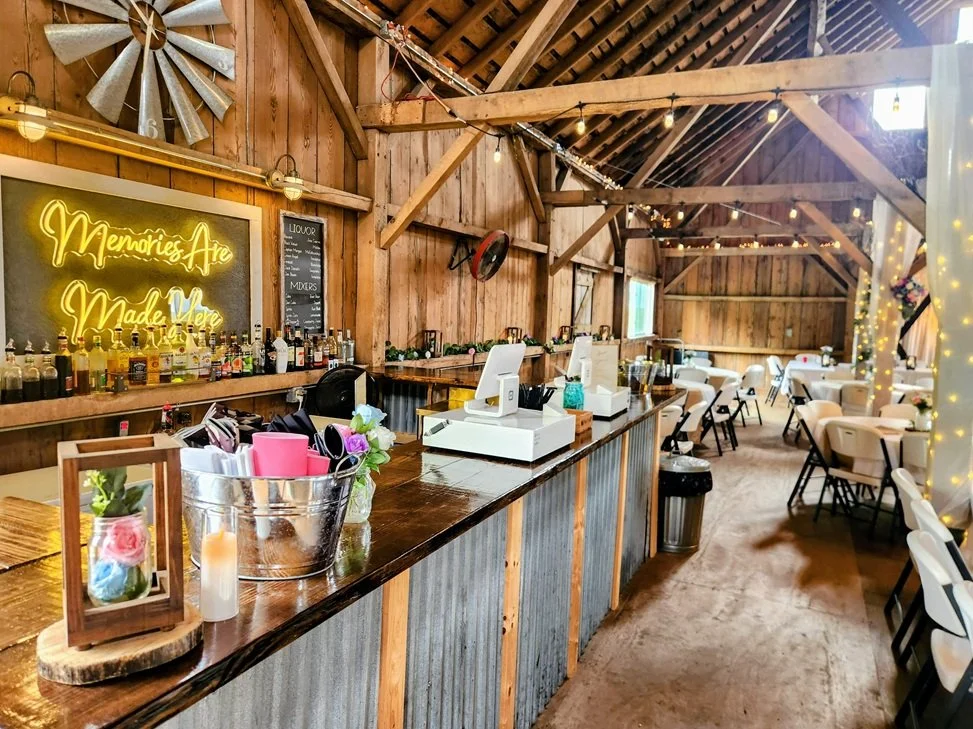 Interior of a rustic event space with a bar area, wooden walls, string lights, and seating. Neon sign reads 'Memories Are Made Here' and bottles of alcohol are lined on the bar counter.
