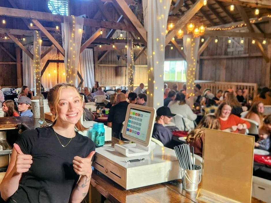Smiling woman giving two thumbs up at rustic event with many people, string lights, and wooden decor.