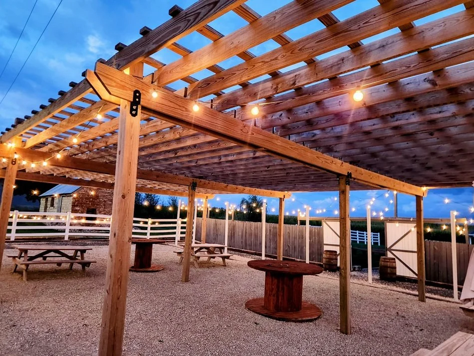 Wooden outdoor patio with string lights, picnic tables, and a gravel ground under a partially open wooden pergola at dusk.