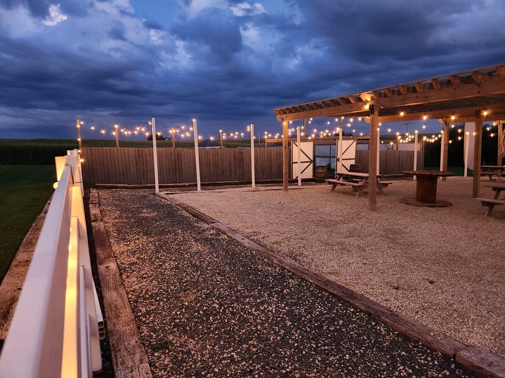 An outdoor patio area decorated with string lights under a cloudy evening sky. The patio has a gravel floor, wooden fences, and a covered seating area with picnic tables.