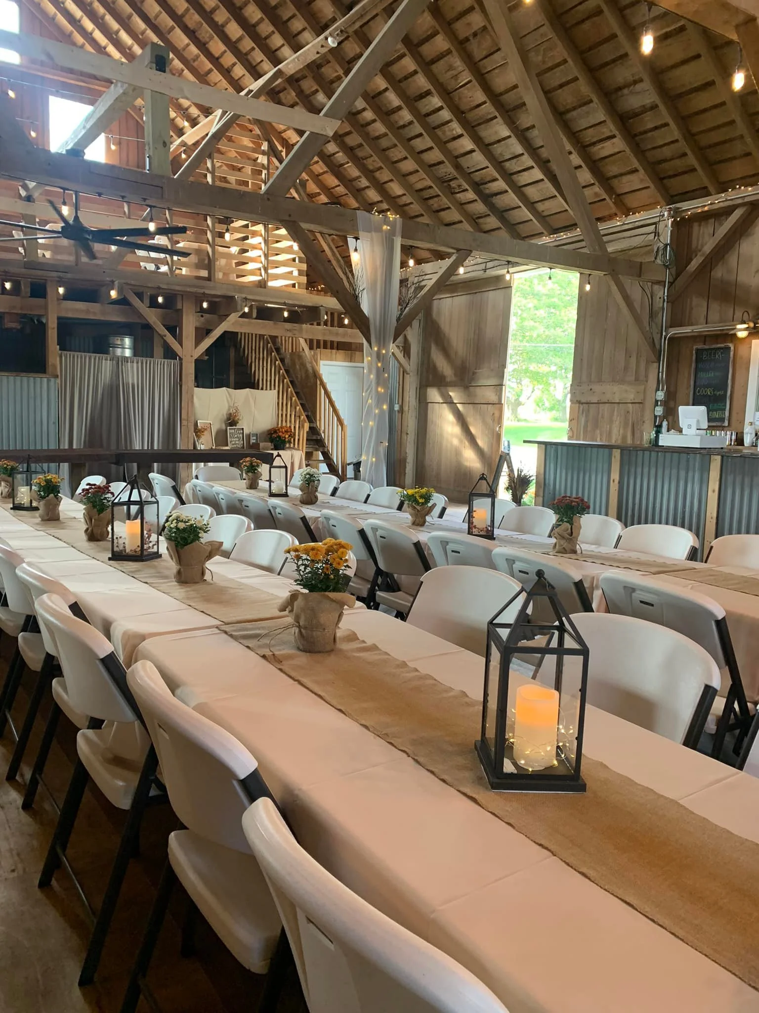 Indoor rustic event space with long dining tables decorated with burlap runners, small potted flowers, candle lanterns, and string lights in a barn with wooden beams and large door opening to greenery outside.