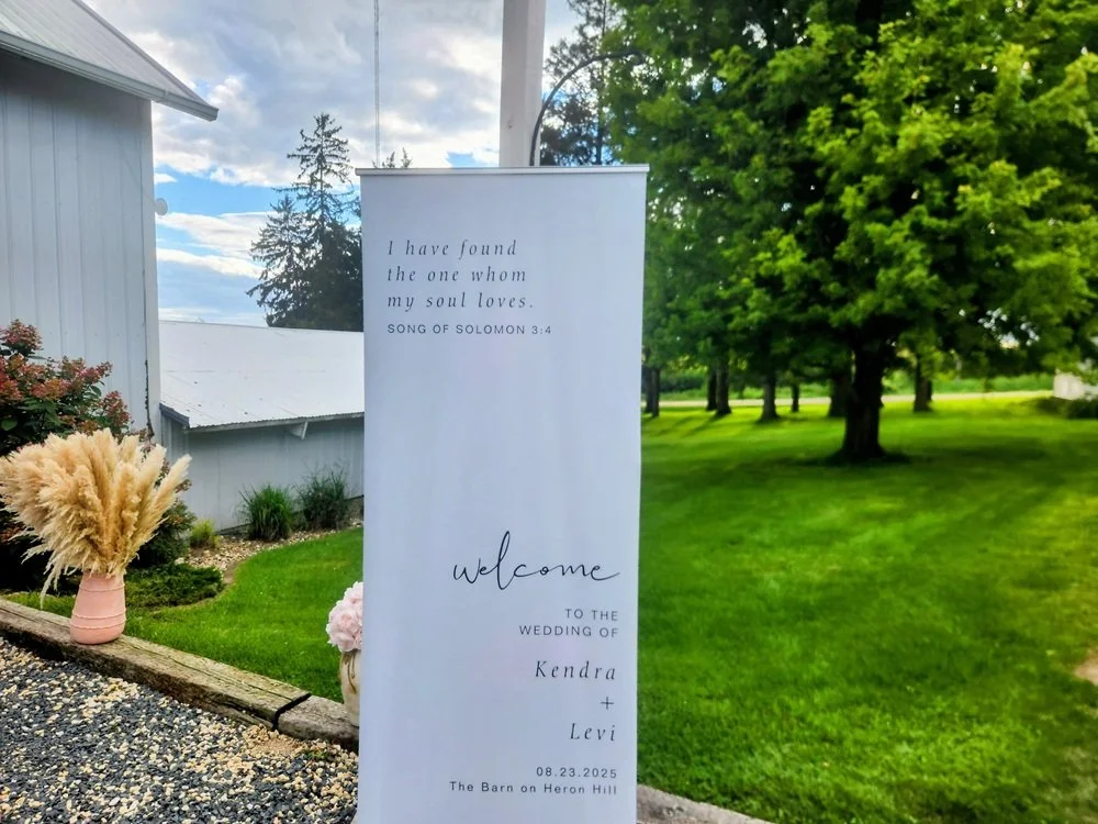 A wedding welcome sign outdoors next to a house and lush green lawn, with trees in the background. The sign features a Bible verse and information about the wedding of Kendra and Levi on August 23, 2025.