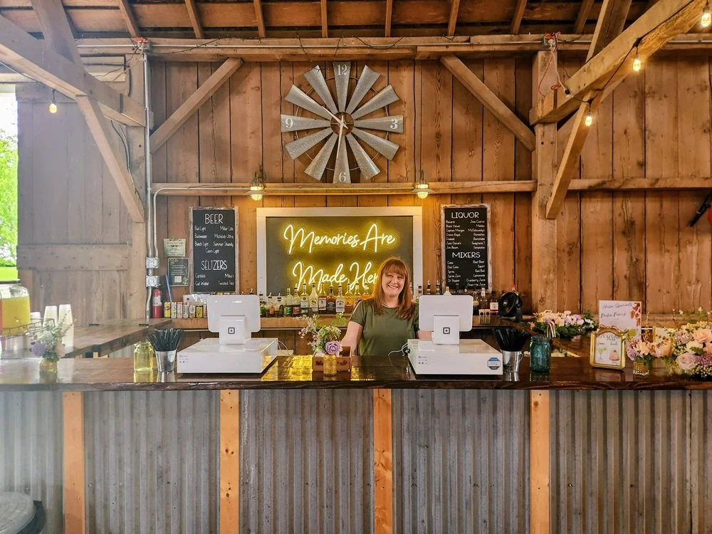 A smiling woman standing behind a rustic bar with a neon sign that says 'Memories Are Made Here' in a barn-style setting. The bar has two cash registers, flowers, and various bottles of alcohol. Chalkboards display drink menus, and warm string lights