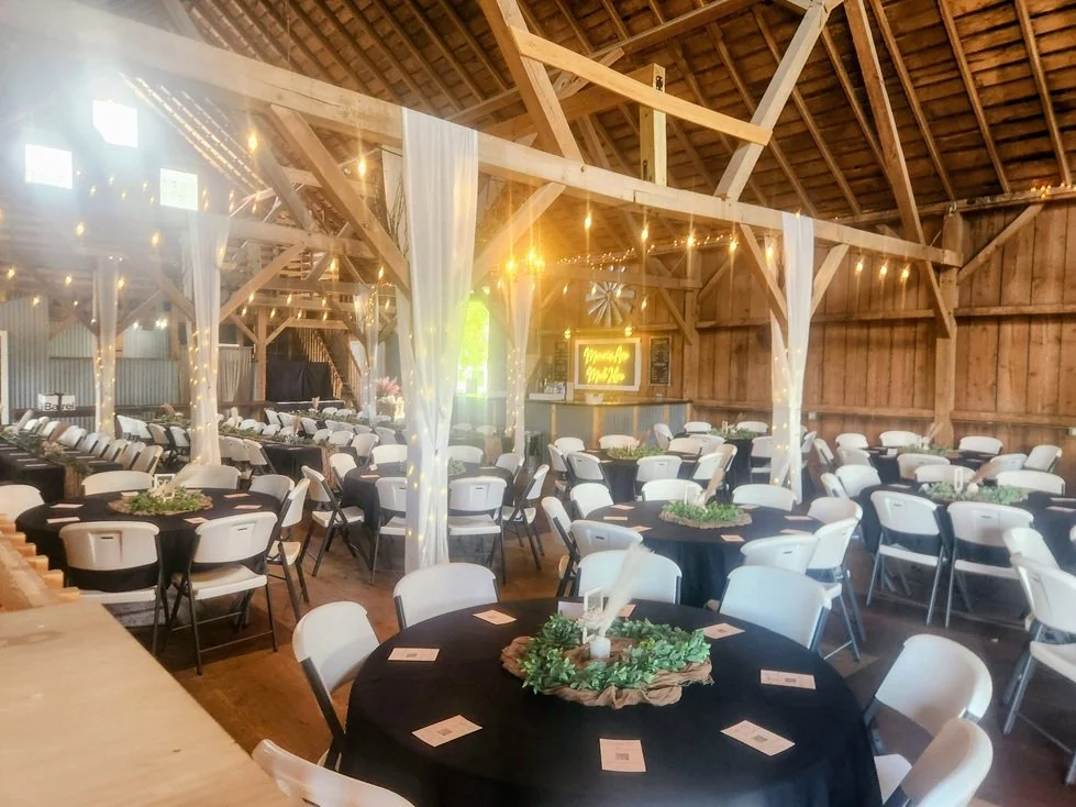 Decorated rustic barn interior set up for a wedding reception with round tables covered in black tablecloths, white chairs, fairy lights, and seasonal centerpieces.