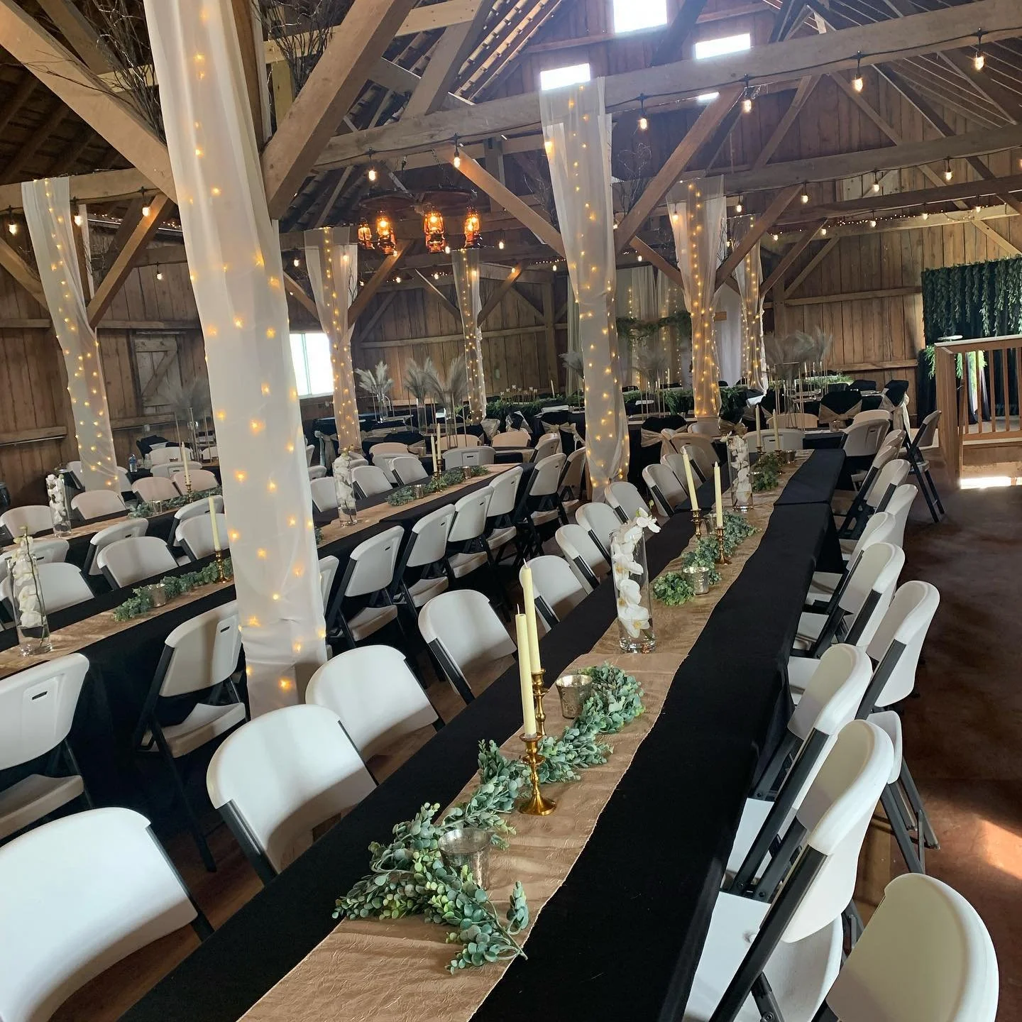 Wedding reception setup inside a rustic wooden barn with long tables decorated with greenery, candles, and white orchids, surrounded by white chairs and draped fairy lights.