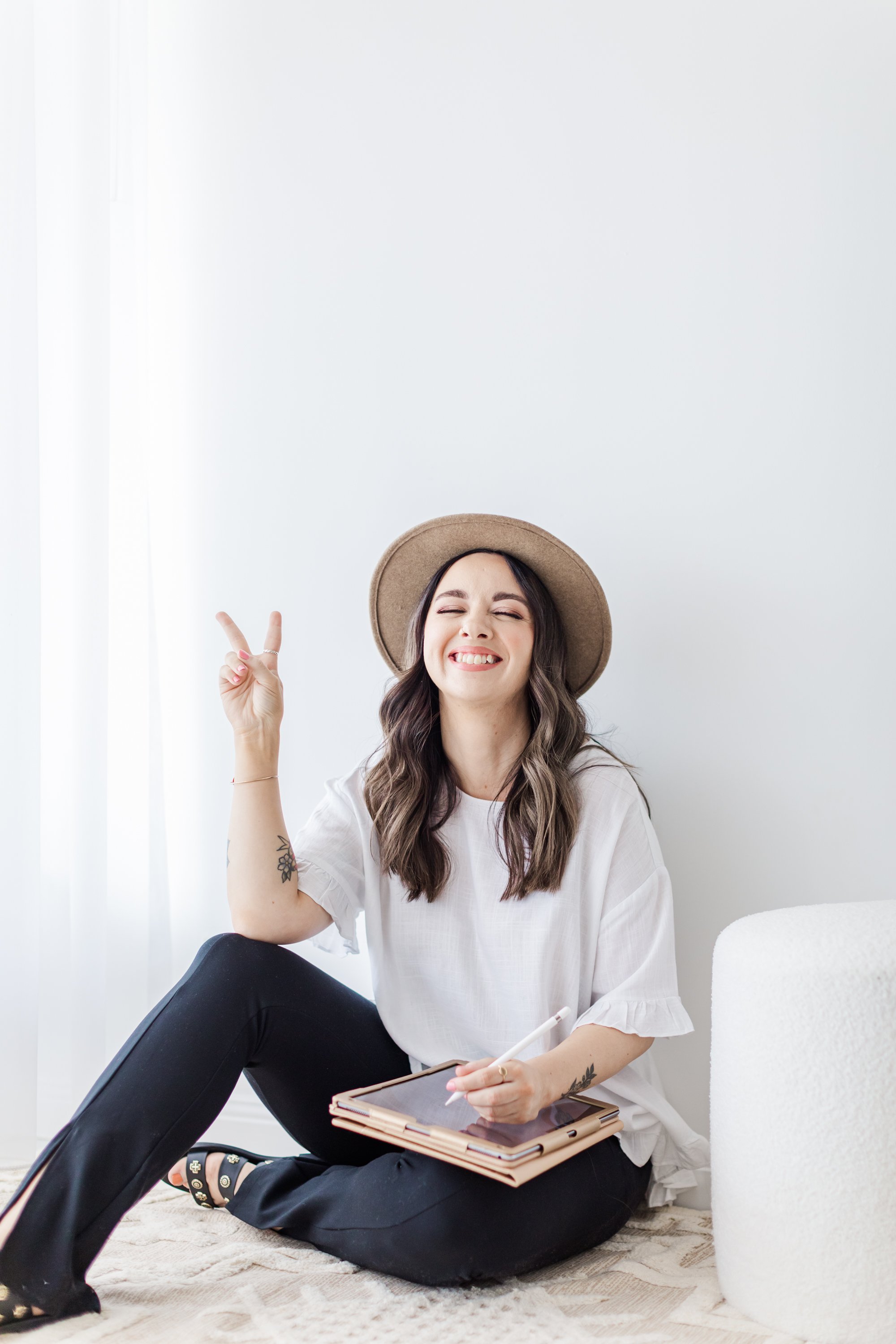 Woman sitting on the floor against a white wall, wearing a hat and smiling. She is holding a tablet and stylus, making a peace sign with her other hand.