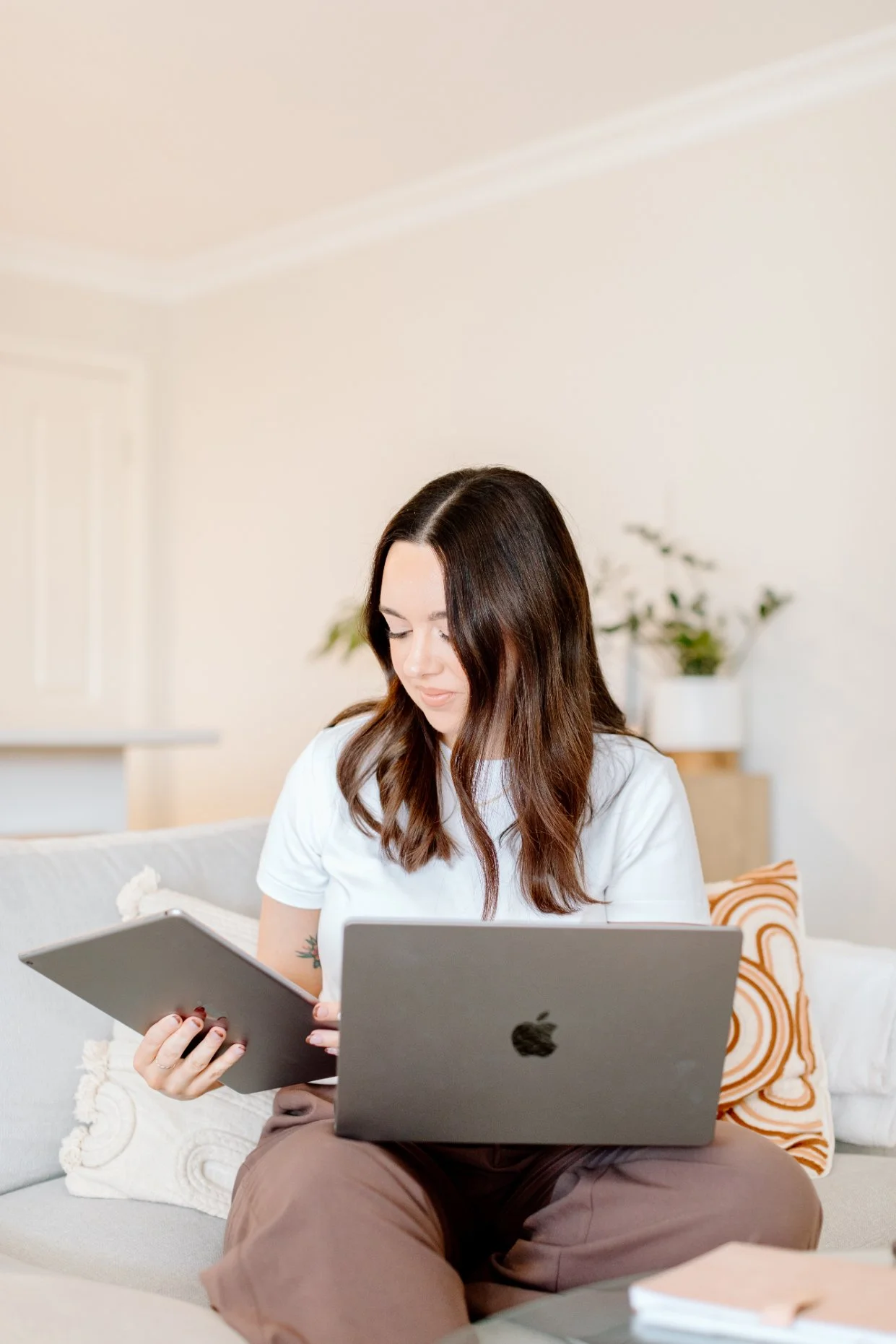 Woman sitting on a couch with a laptop and tablet, wearing a white shirt, with a decorative pillow and plants in the background.