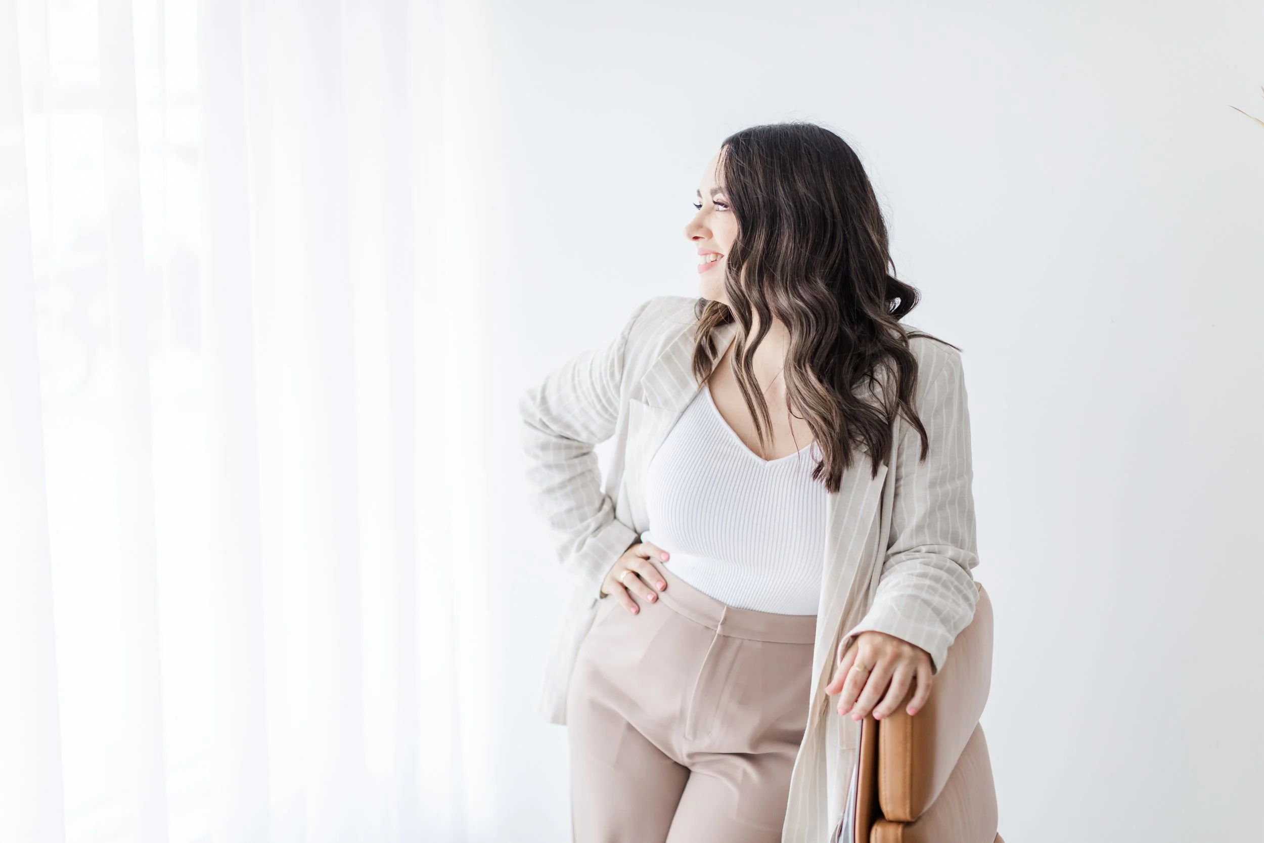 Woman in beige outfit smiling, leaning on chair in bright, minimalistic room.