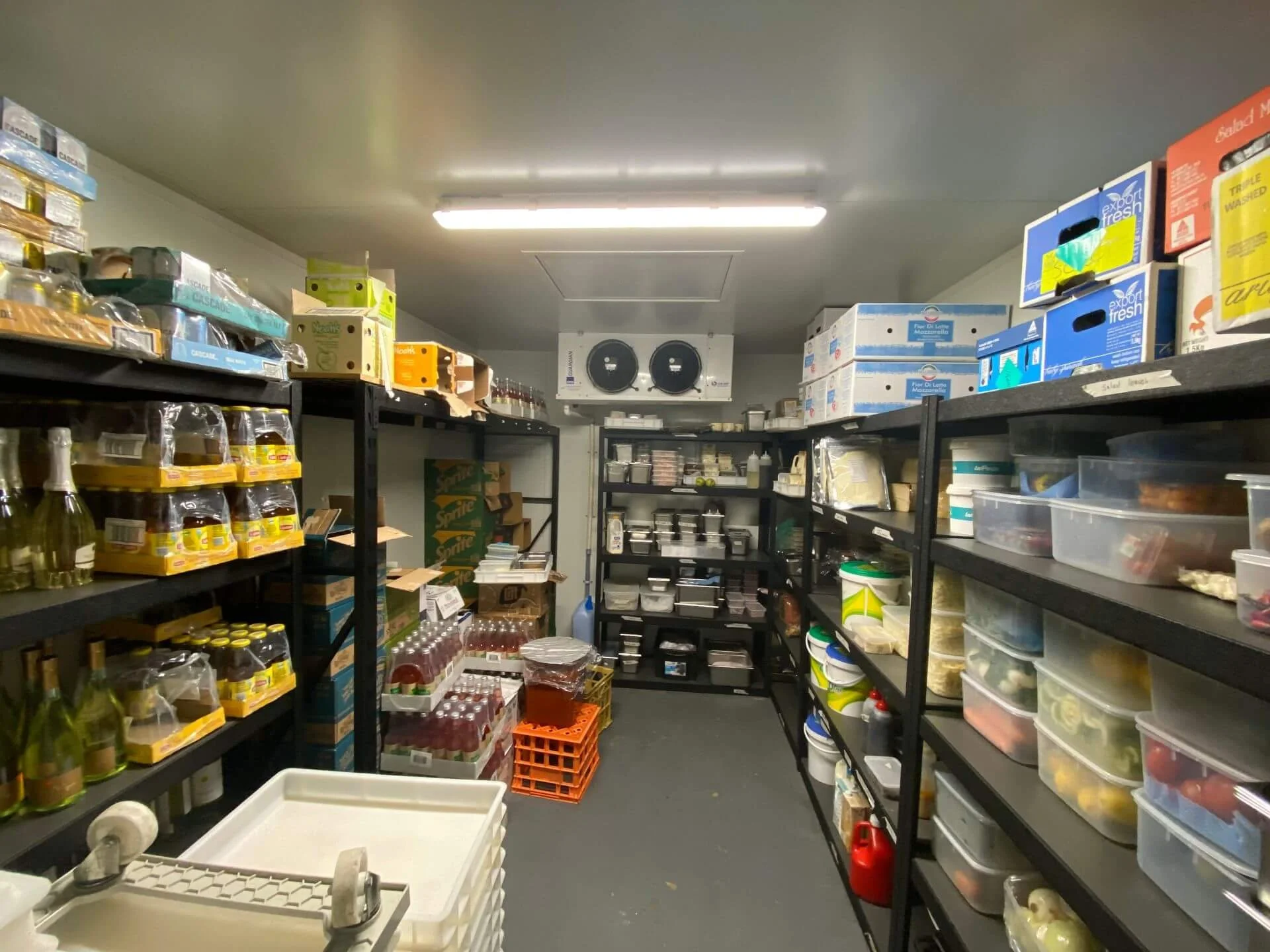 Storage room with black metal shelves filled with food items, containers, and supplies, fluorescent ceiling lights, and a white ceiling.