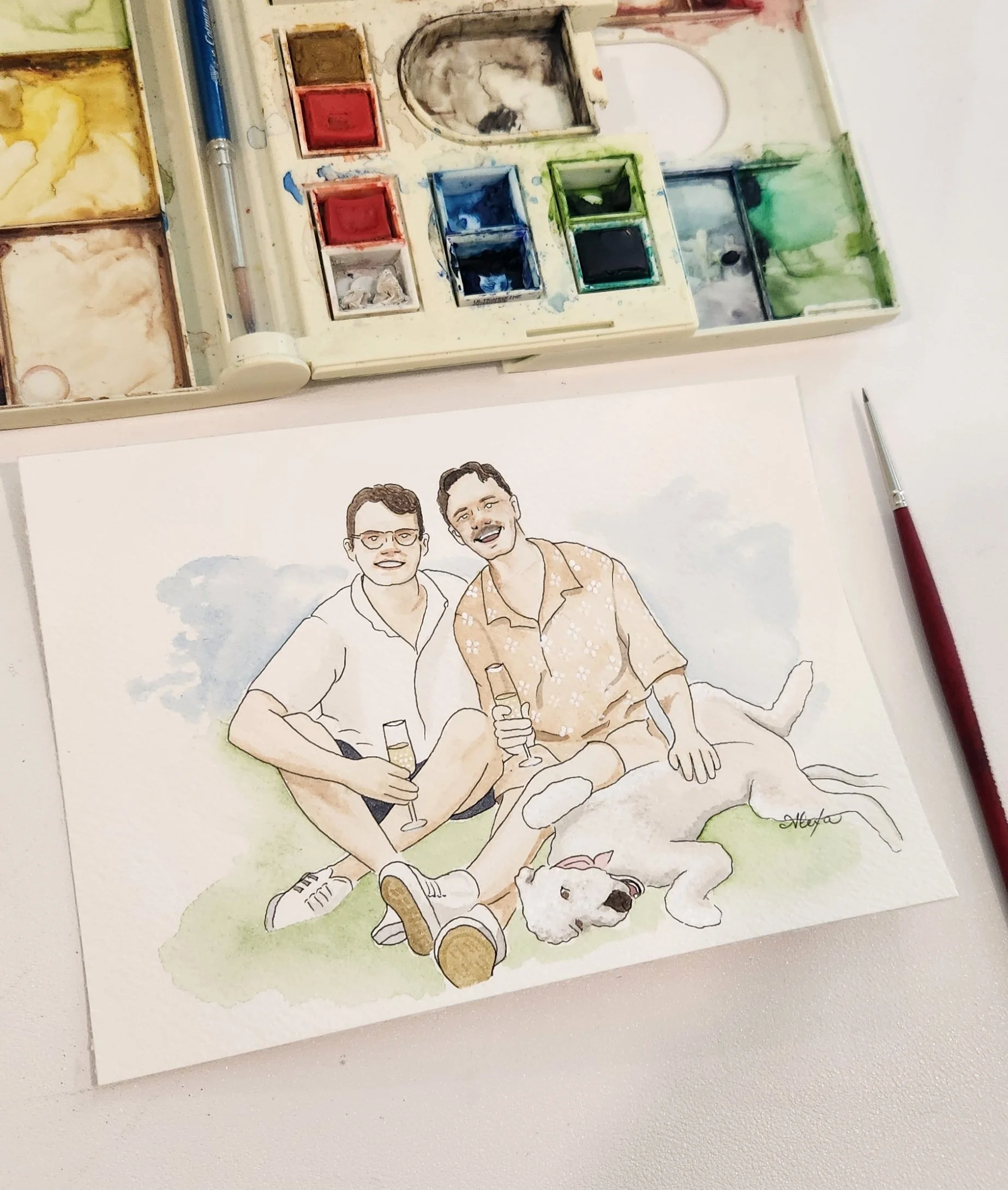 A watercolor illustration of two smiling men sitting on the grass with a dog, holding drinks, with an artist's palette and watercolor brushes nearby. Watercolor engagement portrait by Oklahoma wedding artist Alexa's Illustrations.