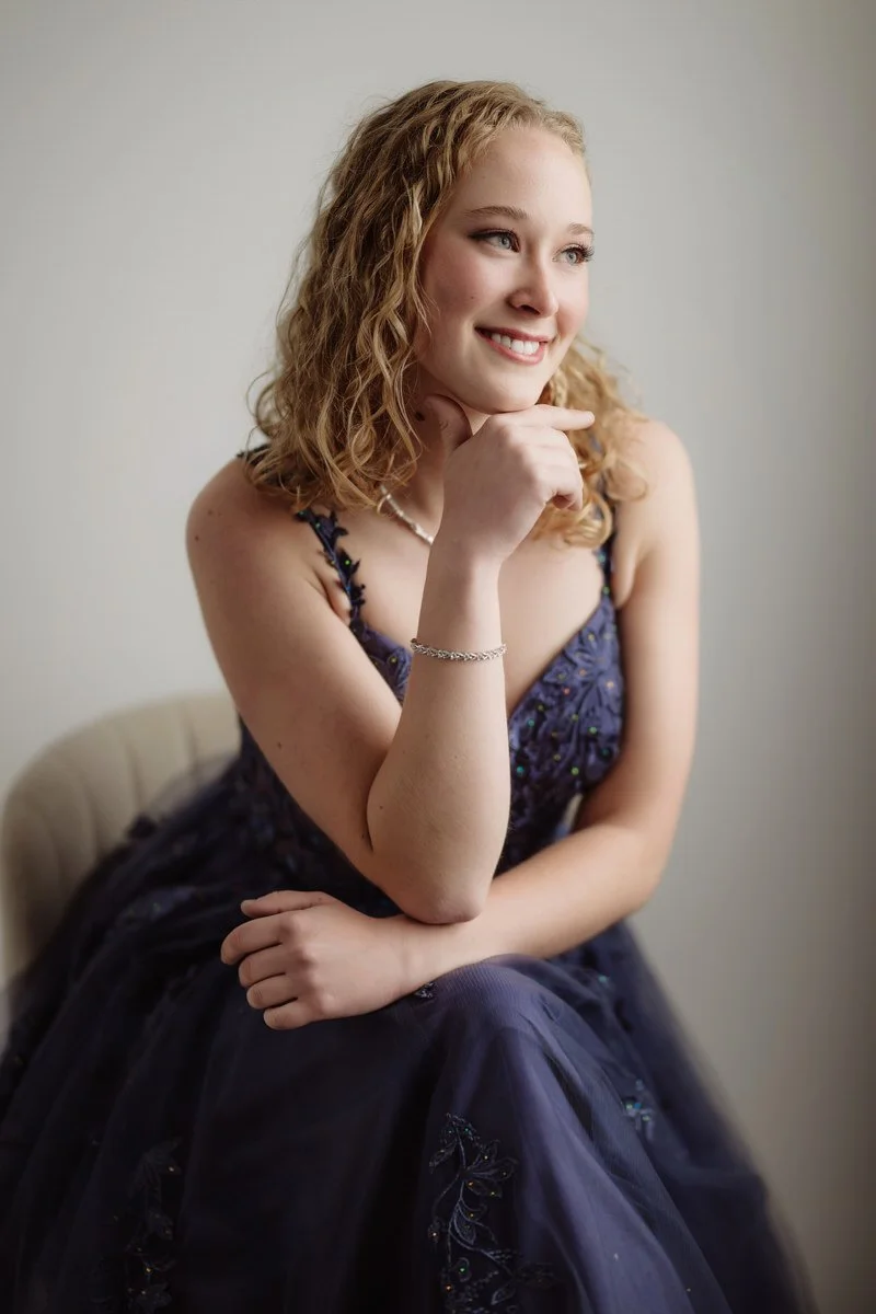 High School prom portrait of girl sitting on a white chair, with a white background, in a blue gown