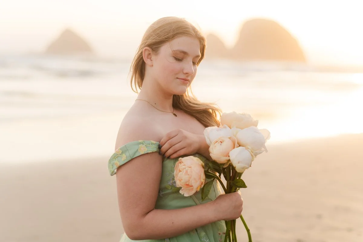 backlight image of high school graduate holding peonies walking along the beach in a green dress