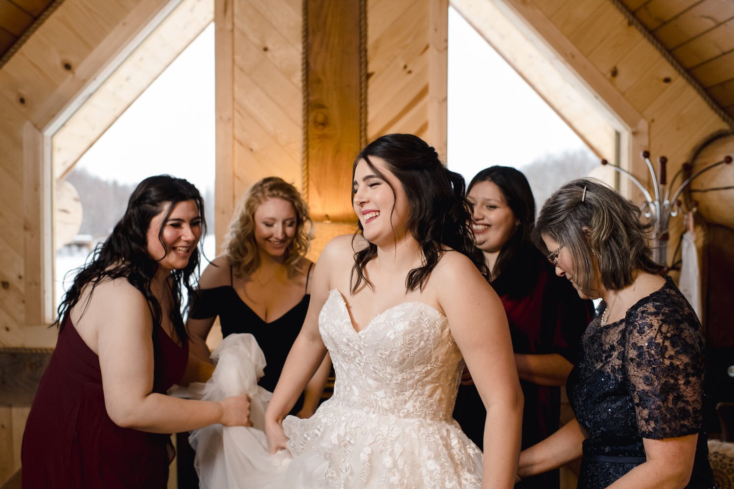 Bride in a white wedding dress surrounded by smiling women helping her get ready, in a wooden room.