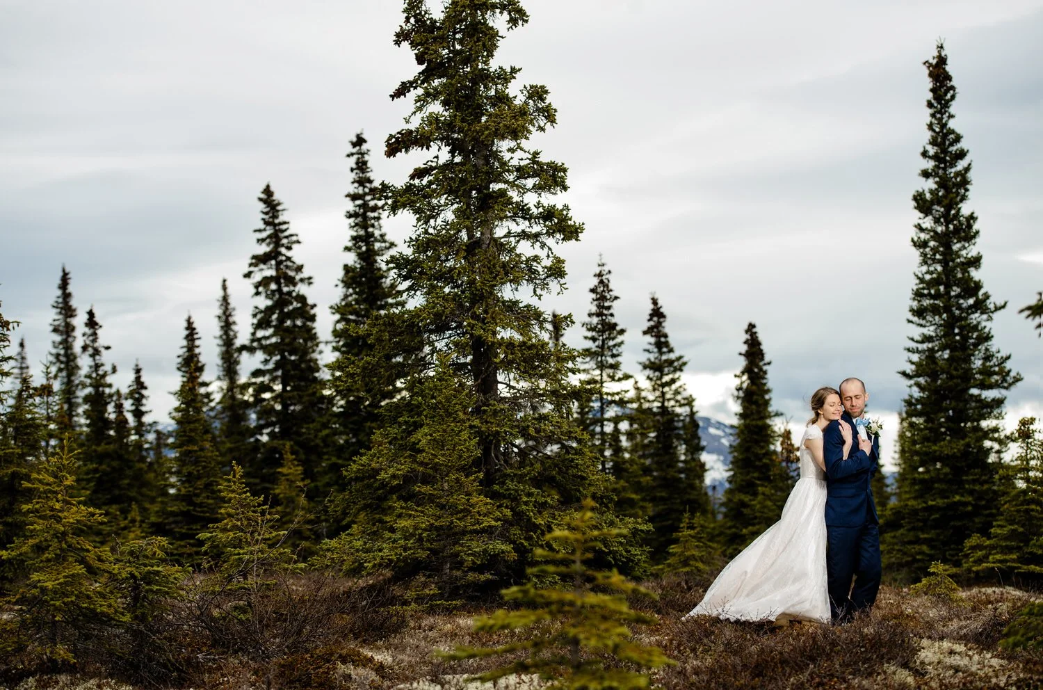 Bride and groom embracing in a forested area, surrounded by tall evergreen trees, with overcast sky and mountains in the background.