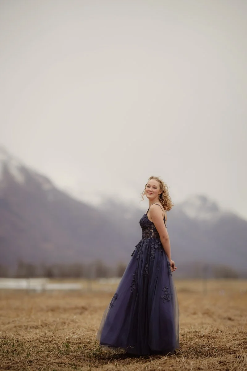 Girl with blonde hair stands with her hands behind her back. Here hair is blowing in the wind for her senior portrait and the mountains in the background.