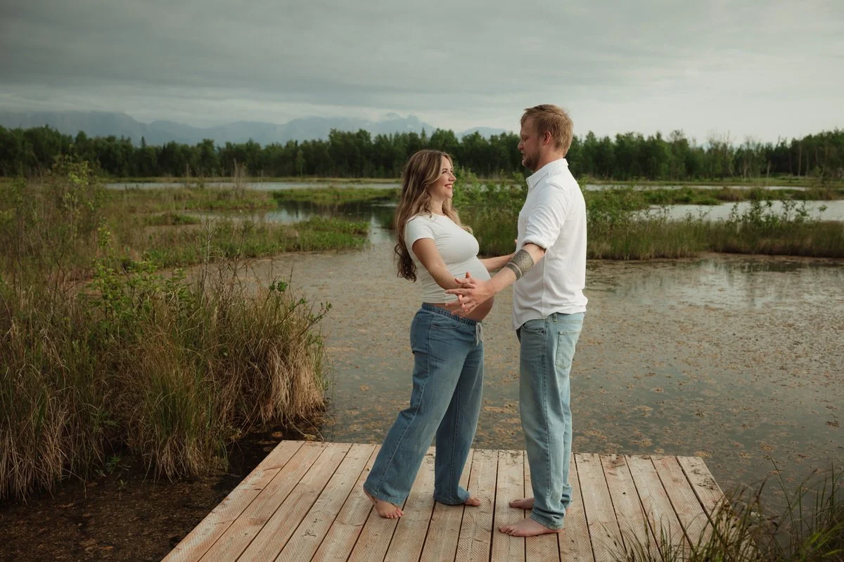 Pregnant couple standing on a dock of a lake holding hands in Palmer, alaska